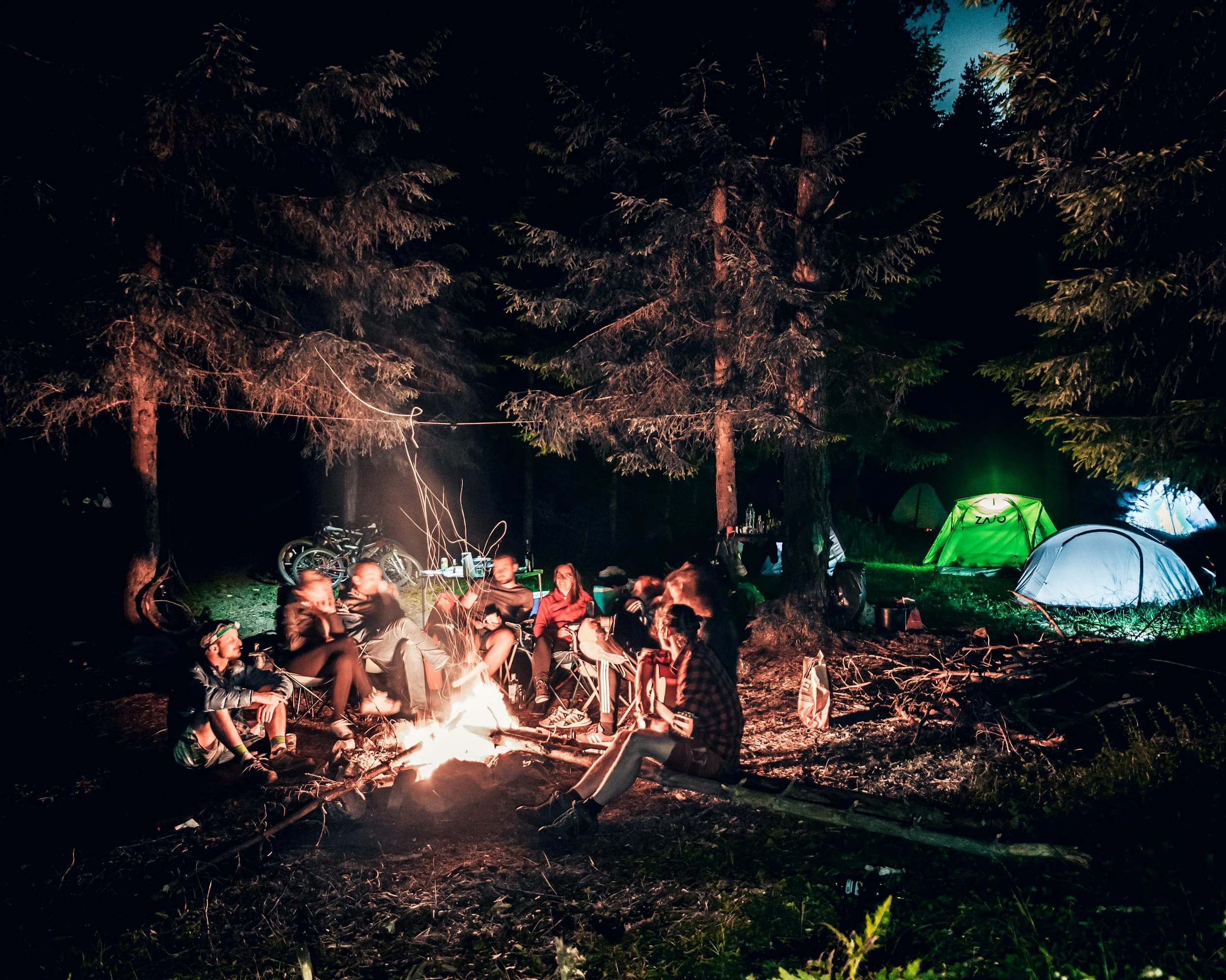 A group of people sitting around a campfire in a forest at night with tents set up and trees in the background.