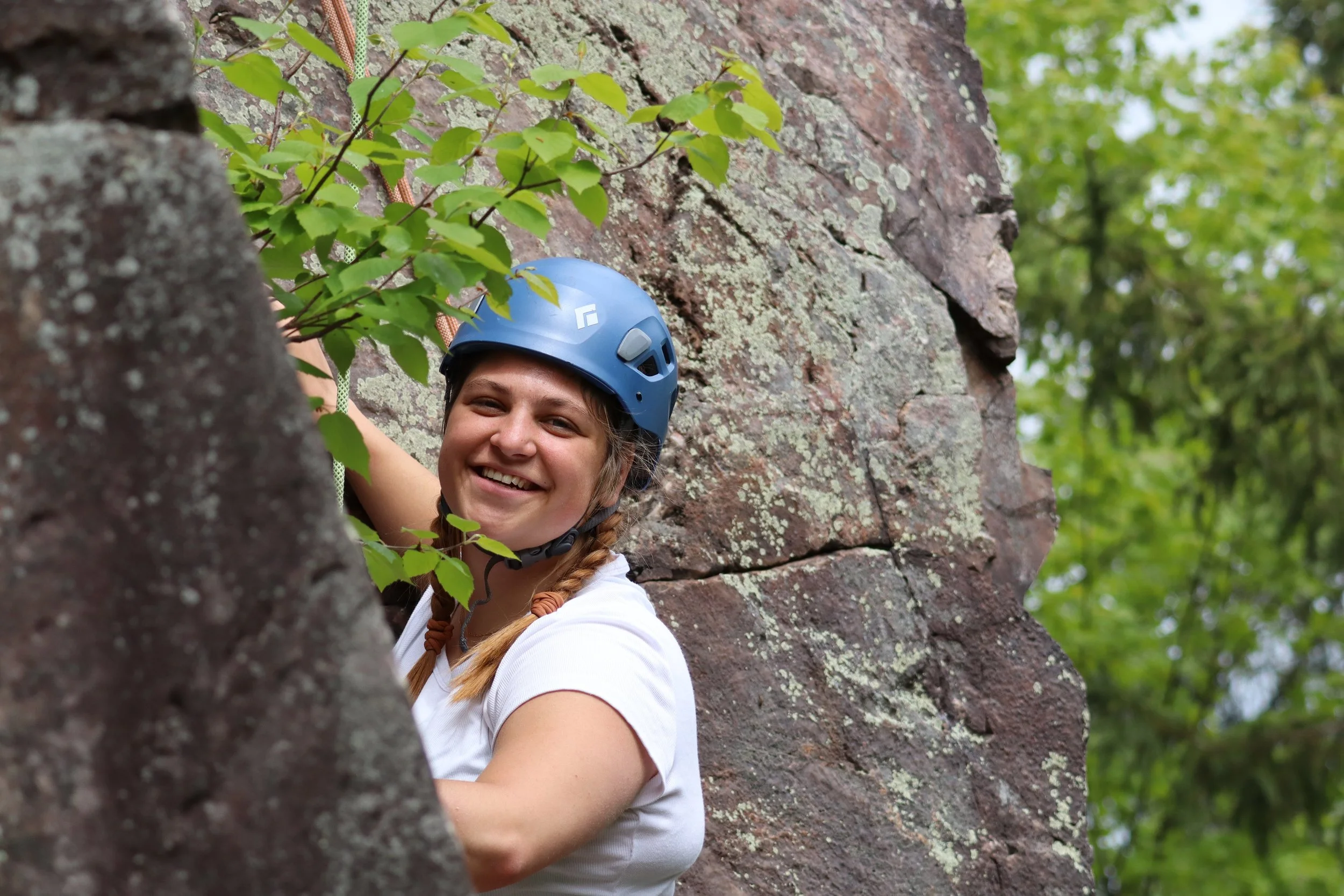 A young woman with braided hair wearing a blue climbing helmet smiling while climbing a rock wall outdoors surrounded by green trees.