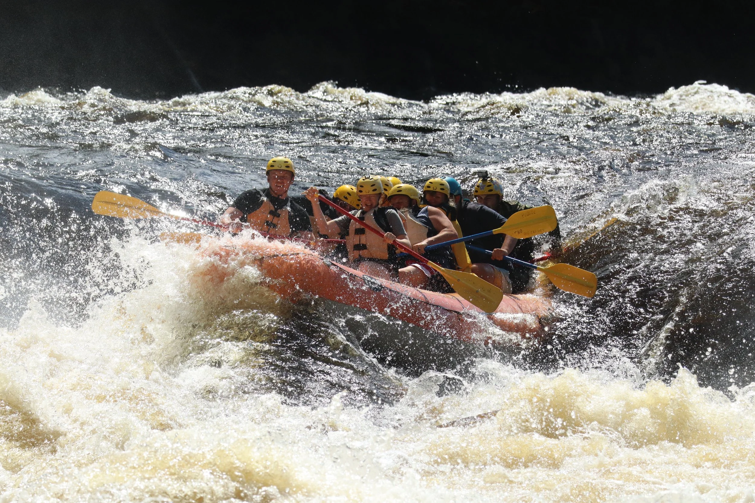 A group of seven people wearing helmets and life jackets rafting through rough water in a canyon.