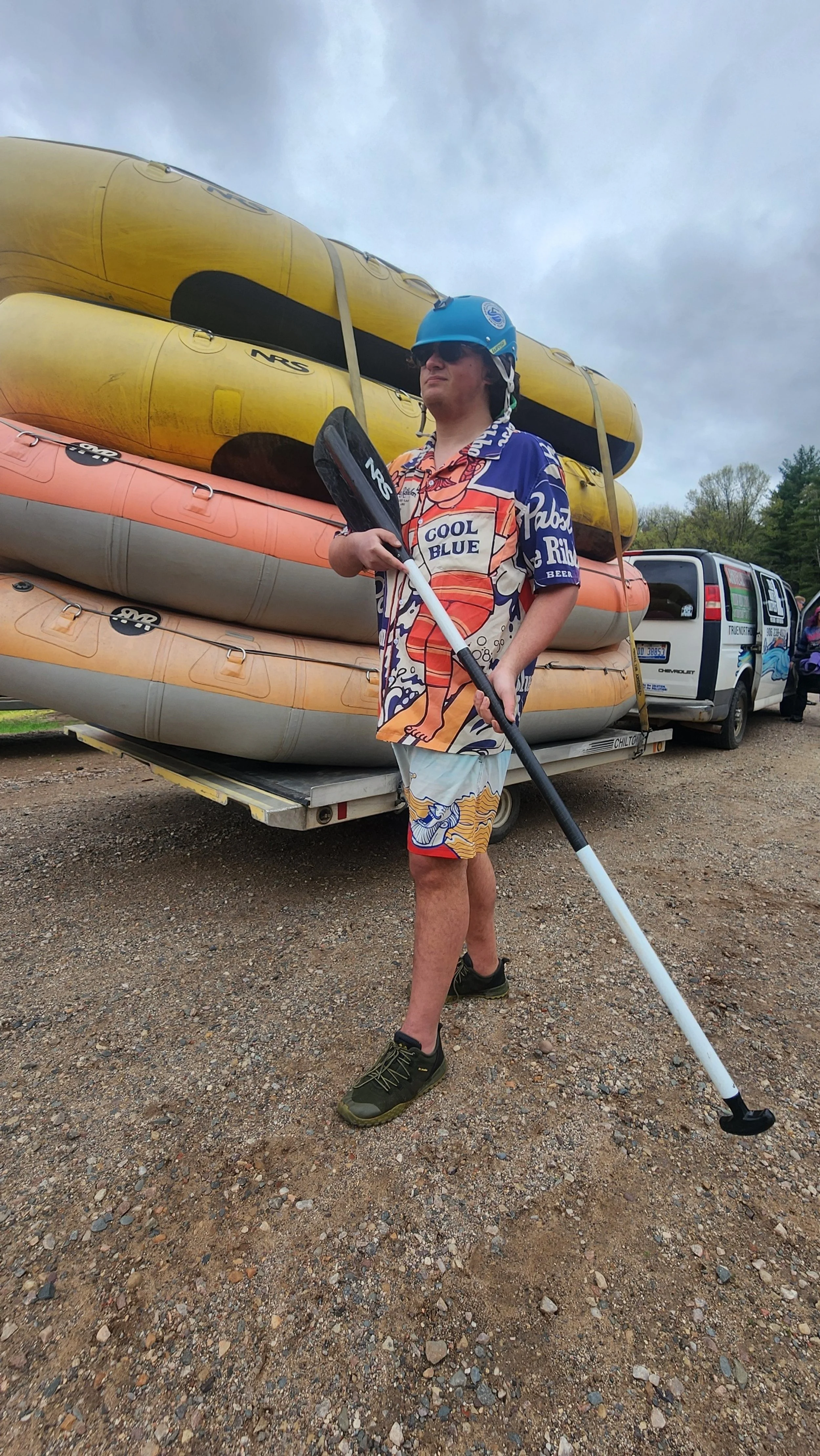 Man wearing sunglasses, blue helmet, and colorful Hawaiian shirt holding a paddle standing in front of a trailer loaded with yellow and orange kayaks, with a van and trees in the background.