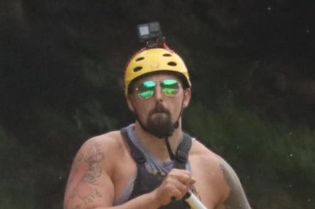 Group of five people white water rafting on a river, wearing helmets and life jackets, surrounded by trees.