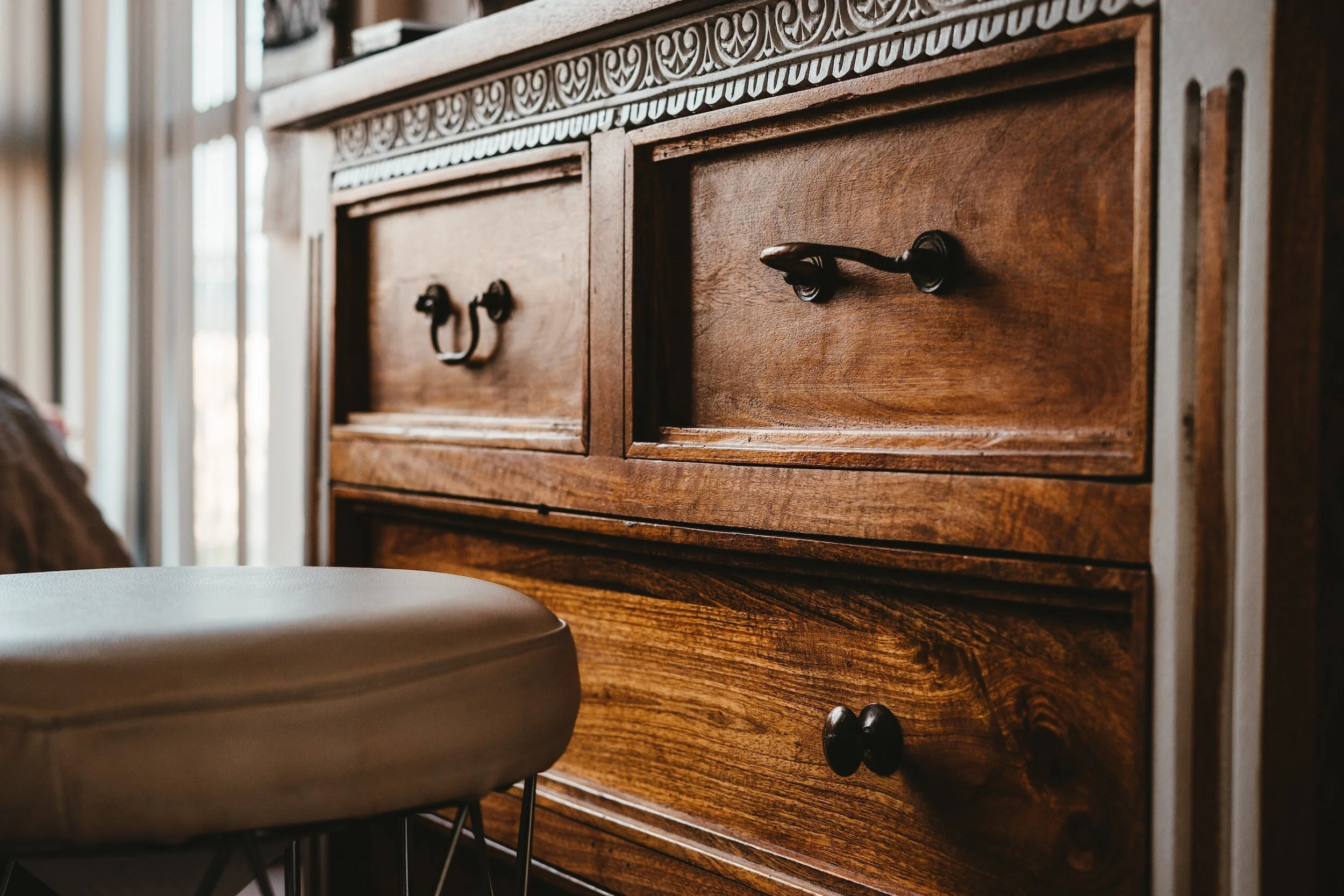 Close-up of a wooden dresser with decorative carvings on top, black handles on the drawers, and a beige cushioned stool in front of it.