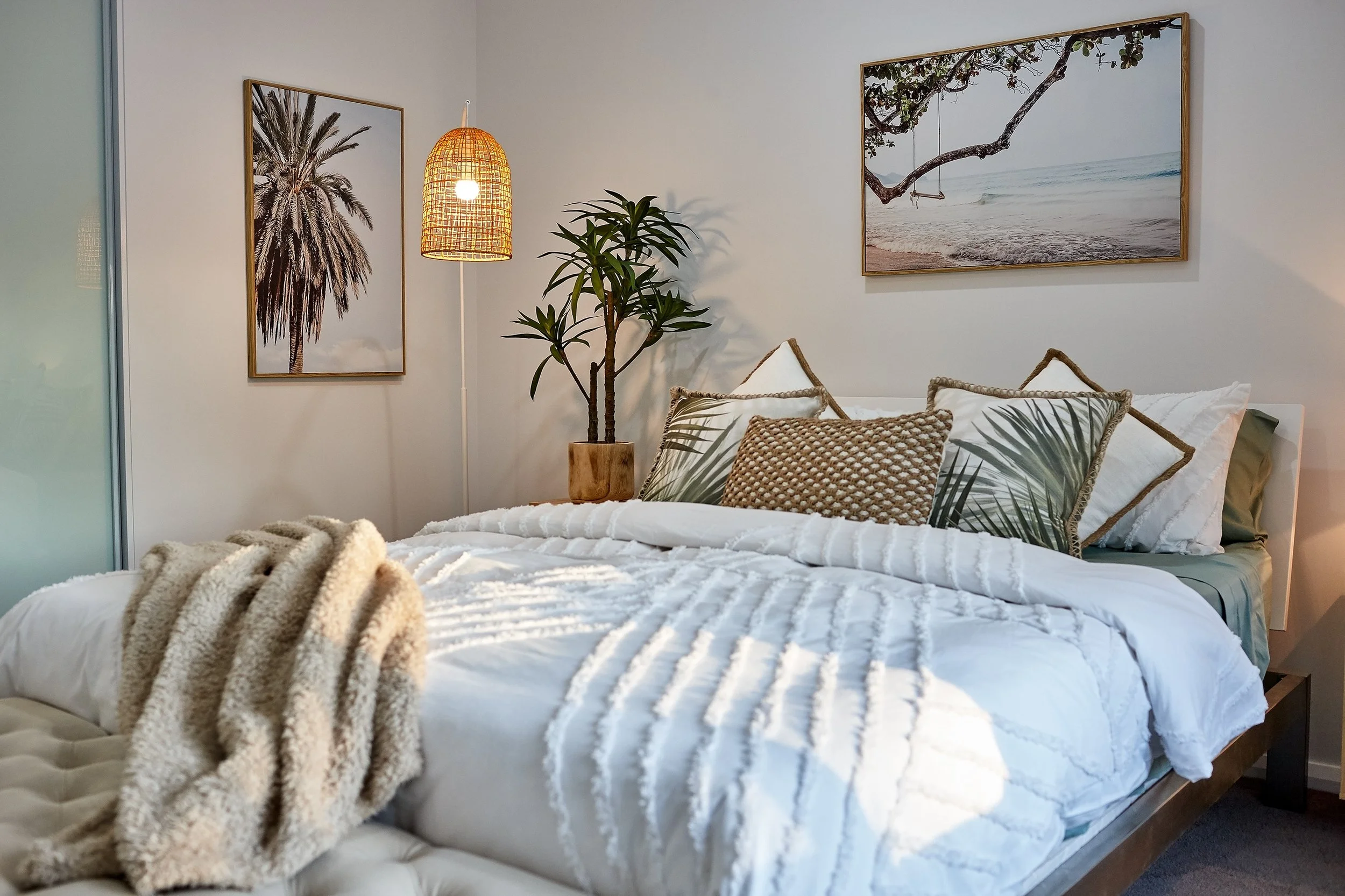 A neatly made bed with white bedding and decorative pillows with nature-inspired designs, a potted plant on the nightstand, framed beach and tree photographs on the wall, and a woven hanging lamp lighting the room