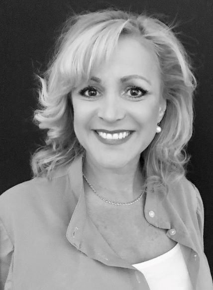 Black and white portrait of a woman with wavy blonde hair, smiling, wearing a light-colored blouse or jacket, pearl earrings, and a necklace against a dark background.