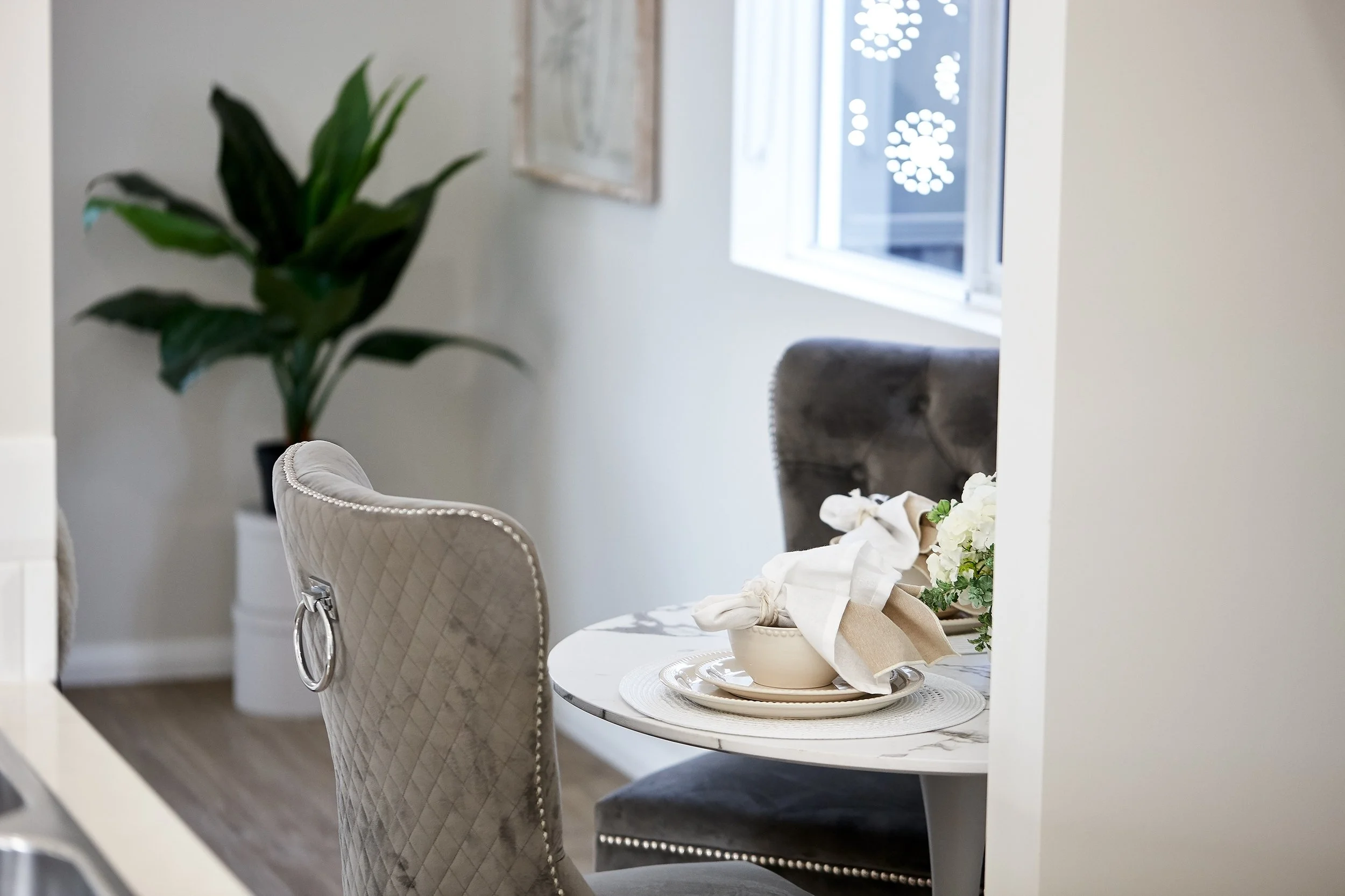 A cozy dining nook with a small round marble table set with a flower arrangement, beige and white plates, napkin, and utensils, surrounded by plush upholstered chairs, next to a window with decorative lights outside and a large potted plant in the background.