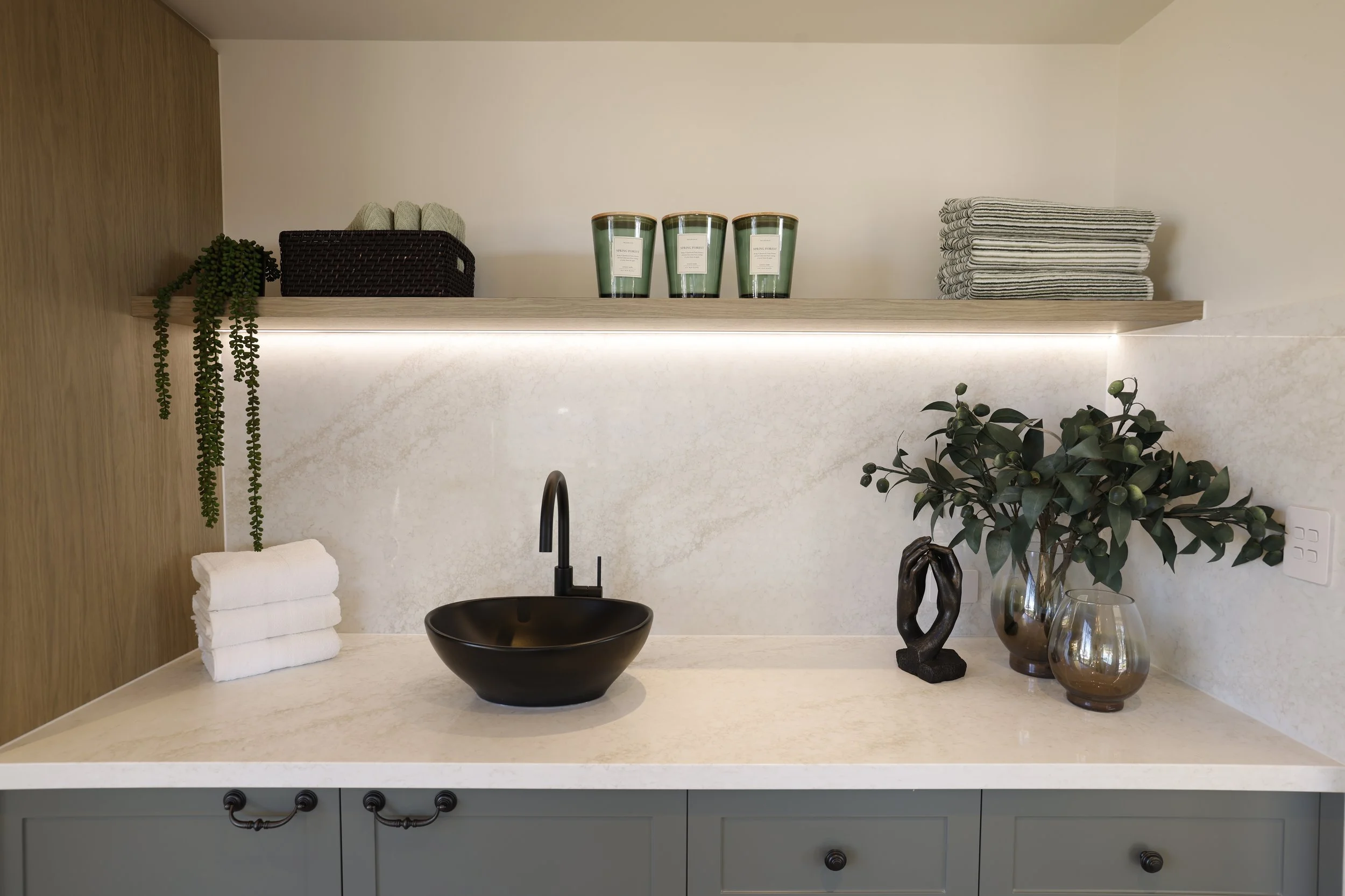 Modern bathroom vanity with black basin sink, black faucet, decorative plants, stacked white towels, and shelves with candles, towels, and a black basket.
