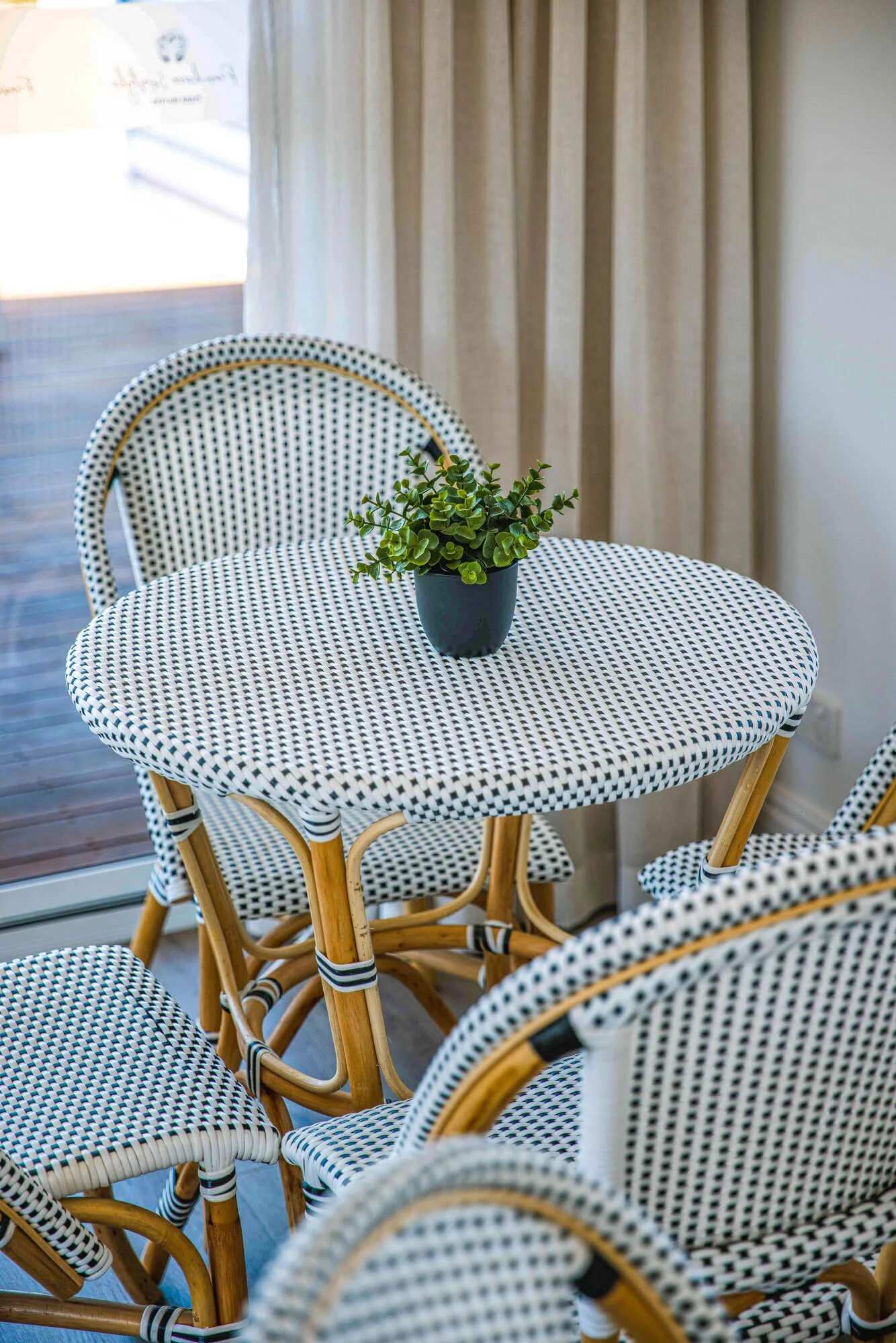 A round table with a black potted plant on top, surrounded by four rattan chairs with black and white polka dot cushions, next to a window with beige curtains.
