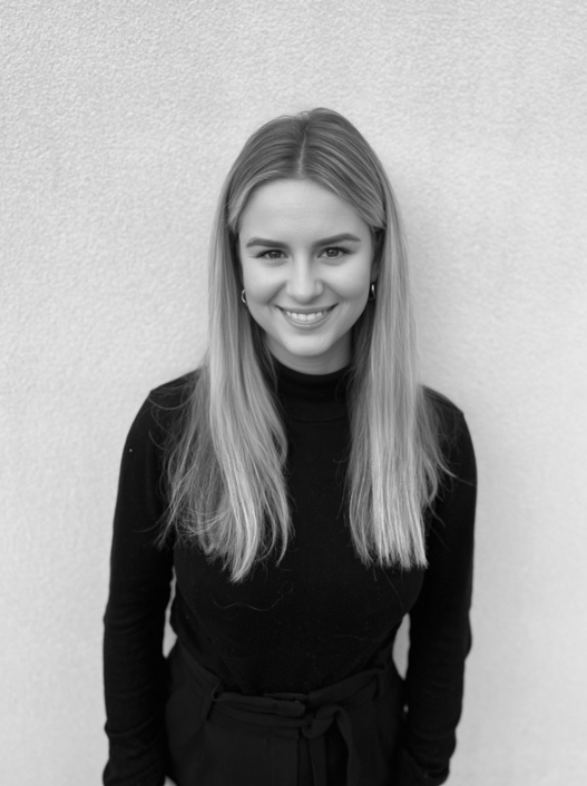 Black and white portrait of a young woman with long, straight hair, smiling at the camera, standing against a plain wall, wearing a black turtleneck and high-waisted pants.
