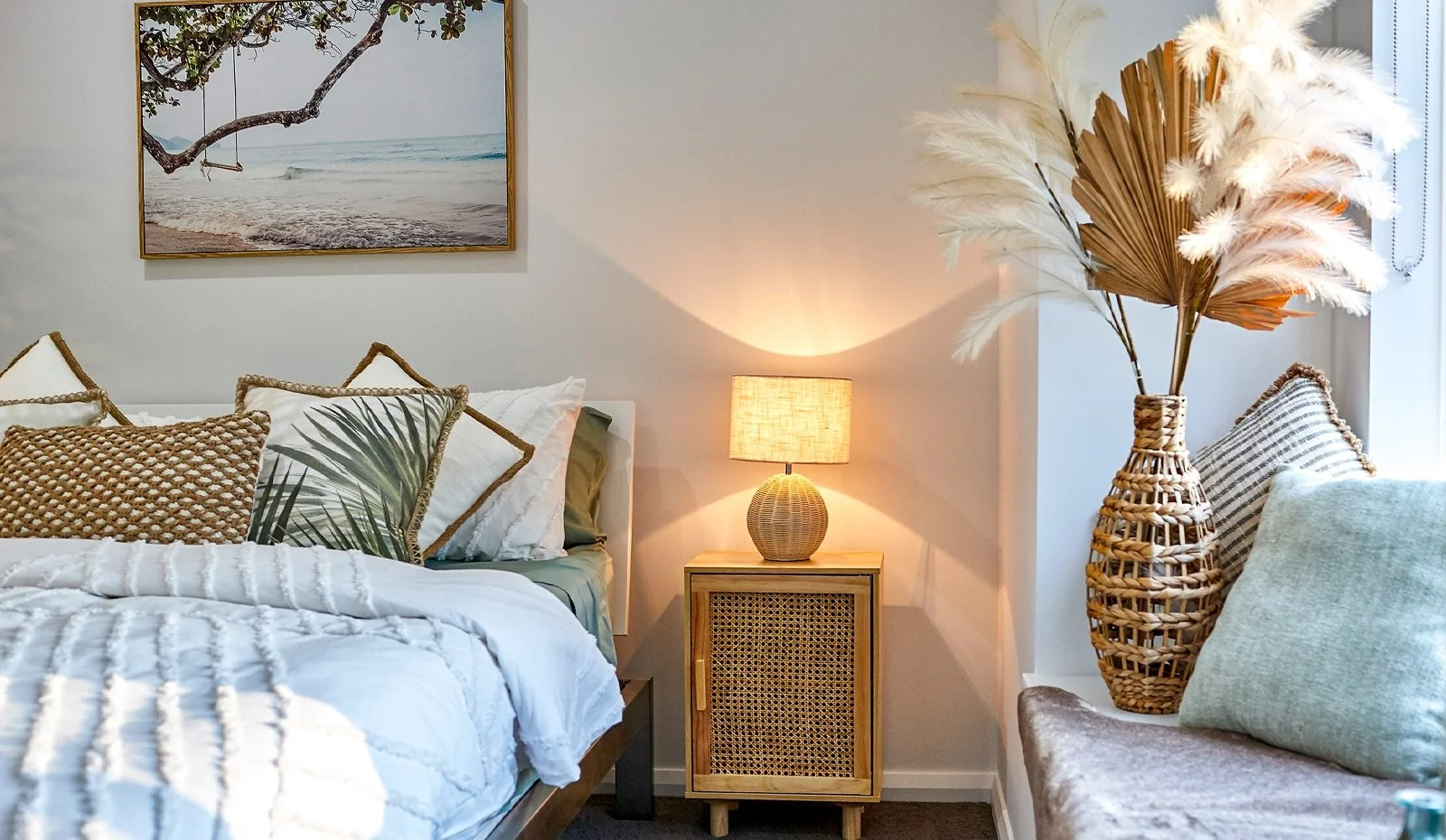 A bedroom with a bed, pillows, a wooden nightstand with a lamp, and a large vase with pampas grass and palm leaves. There is a framed beach scene painting on the wall.
