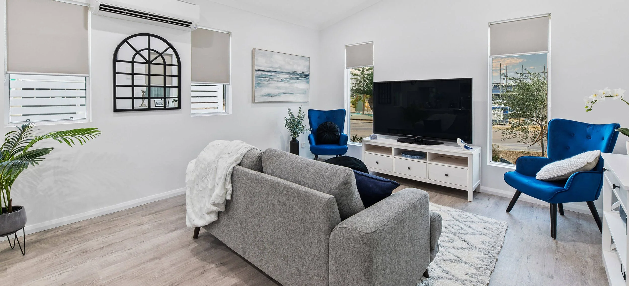 Living room with gray sofa, blue armchairs, a white TV stand with a large flat-screen TV, windows with shades, a plant, and wall art.