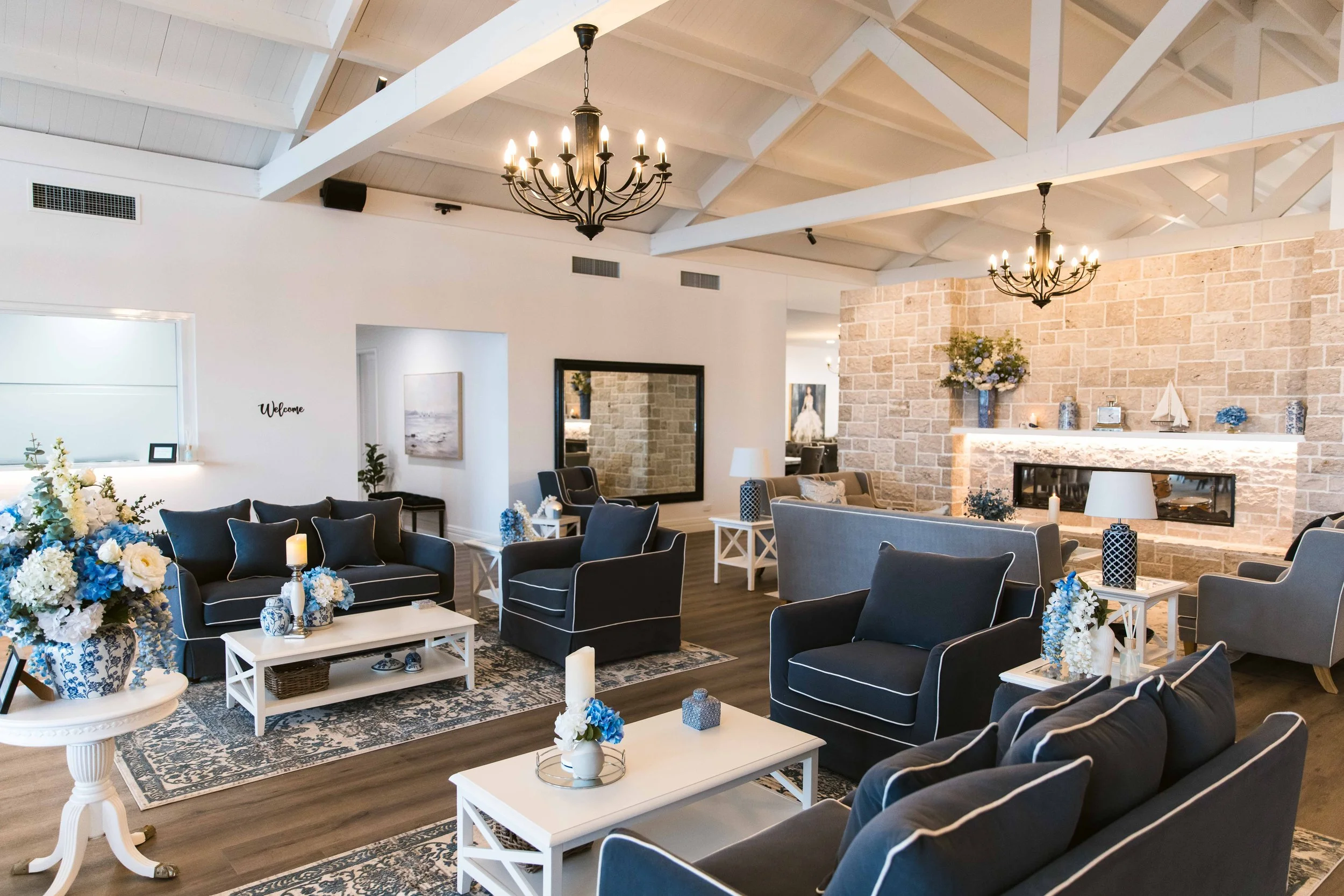A spacious living room or lounge area decorated with navy blue and gray furniture, white side tables, floral arrangements in blue and white vases, a stone fireplace, black chandeliers, and large mirrors and artwork on white walls.