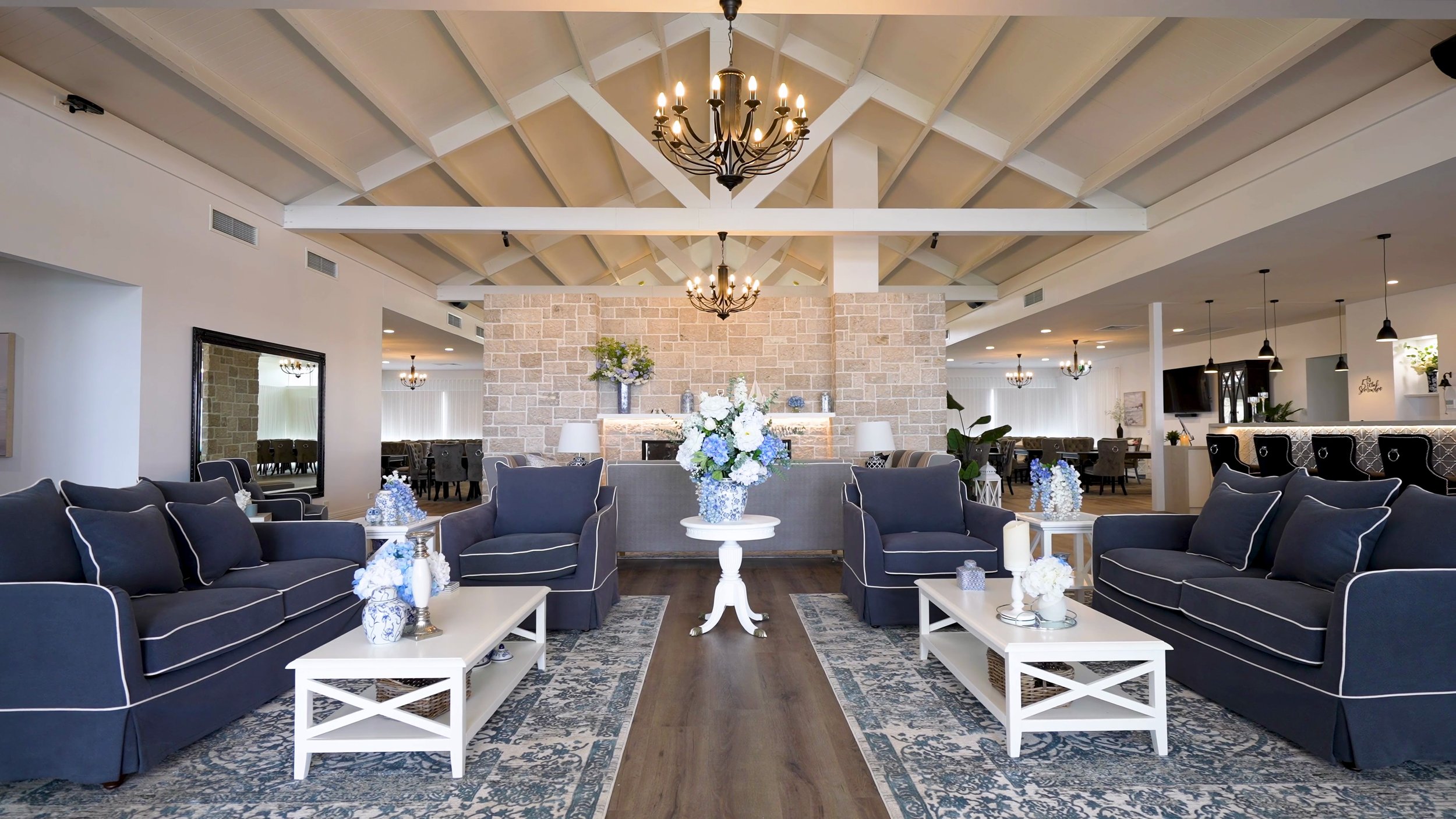 Elegant lounge area with navy blue sofas, white tables, floral centerpieces, and a vaulted ceiling with chandeliers in a modern, well-lit interior.