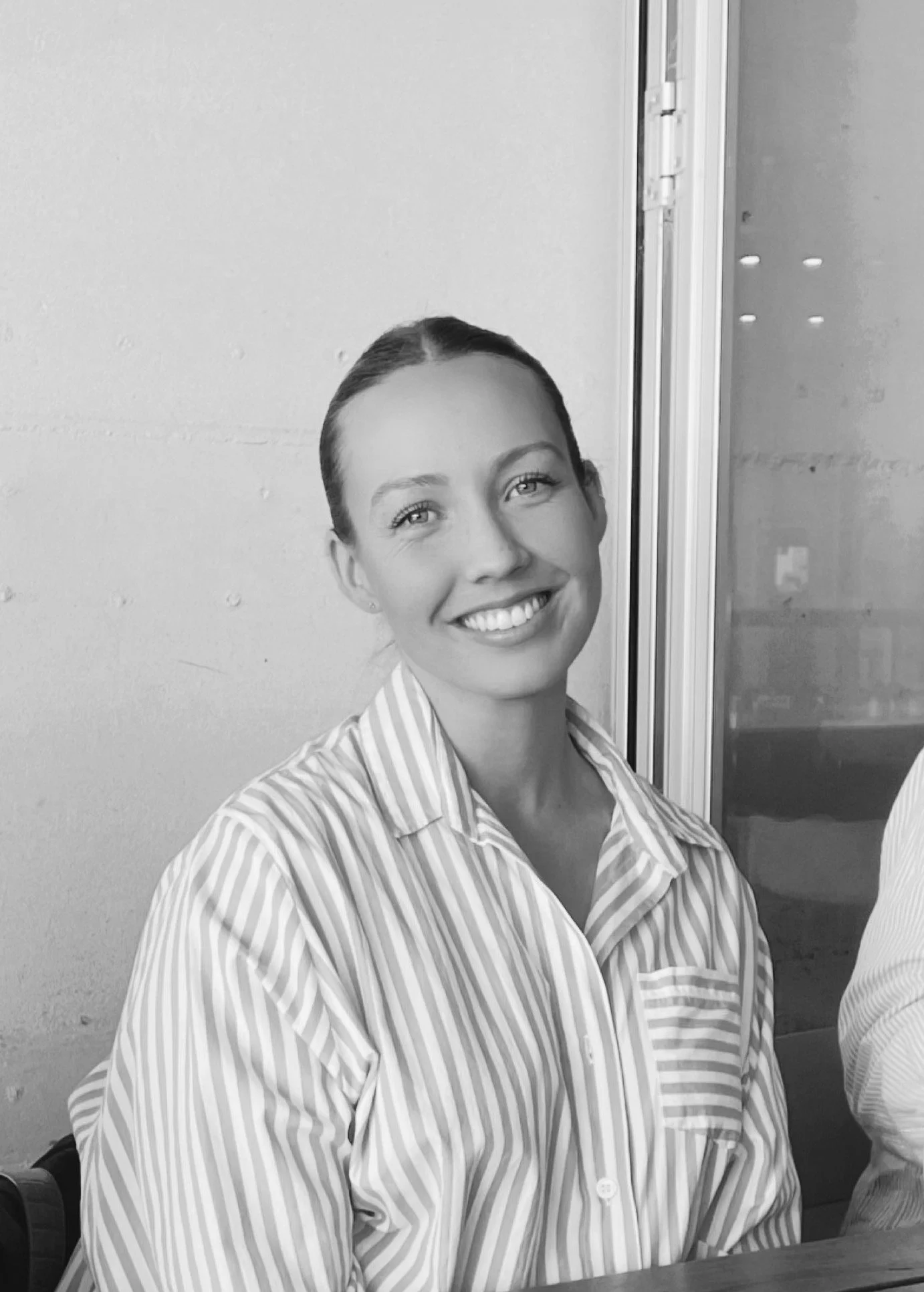 A young woman with dark hair tied back, smiling, wearing a striped button-up shirt, sitting indoors near a glass door.