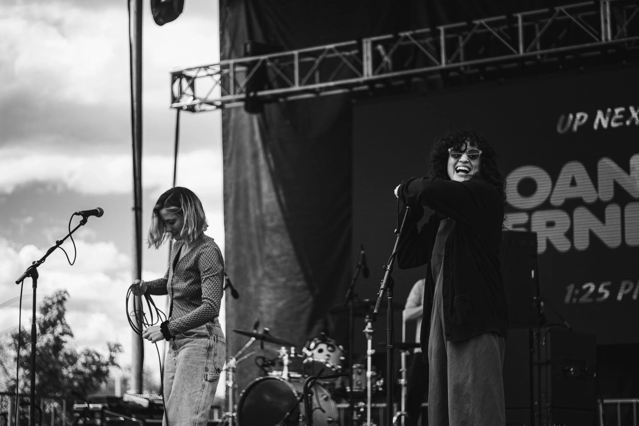 Two female musicians performing on an outdoor stage, one with curly hair and sunglasses singing into a microphone, the other adjusting equipment, with a drum set in the background.
