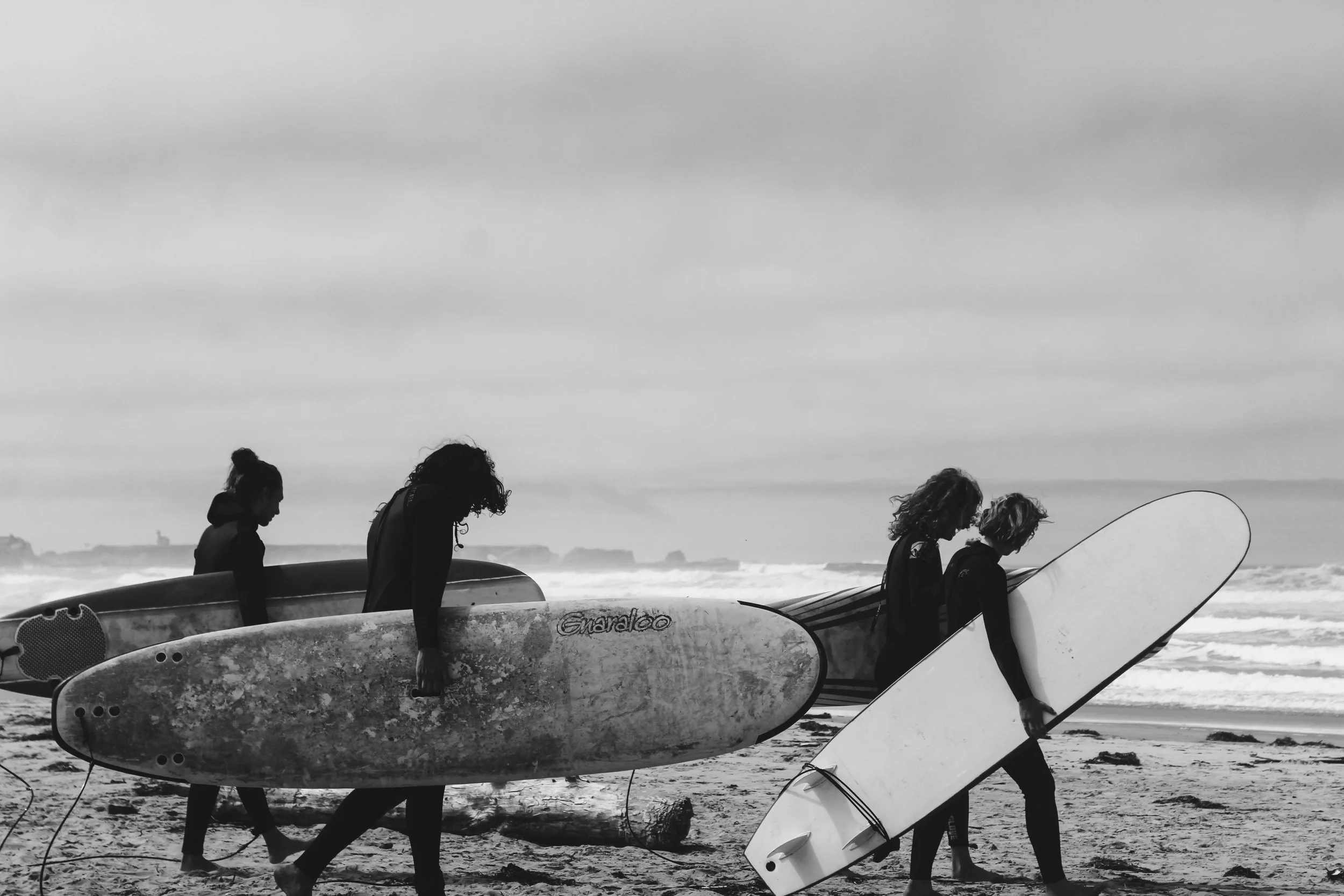 Four people with surfboards walking on a beach with waves and cloudy sky.