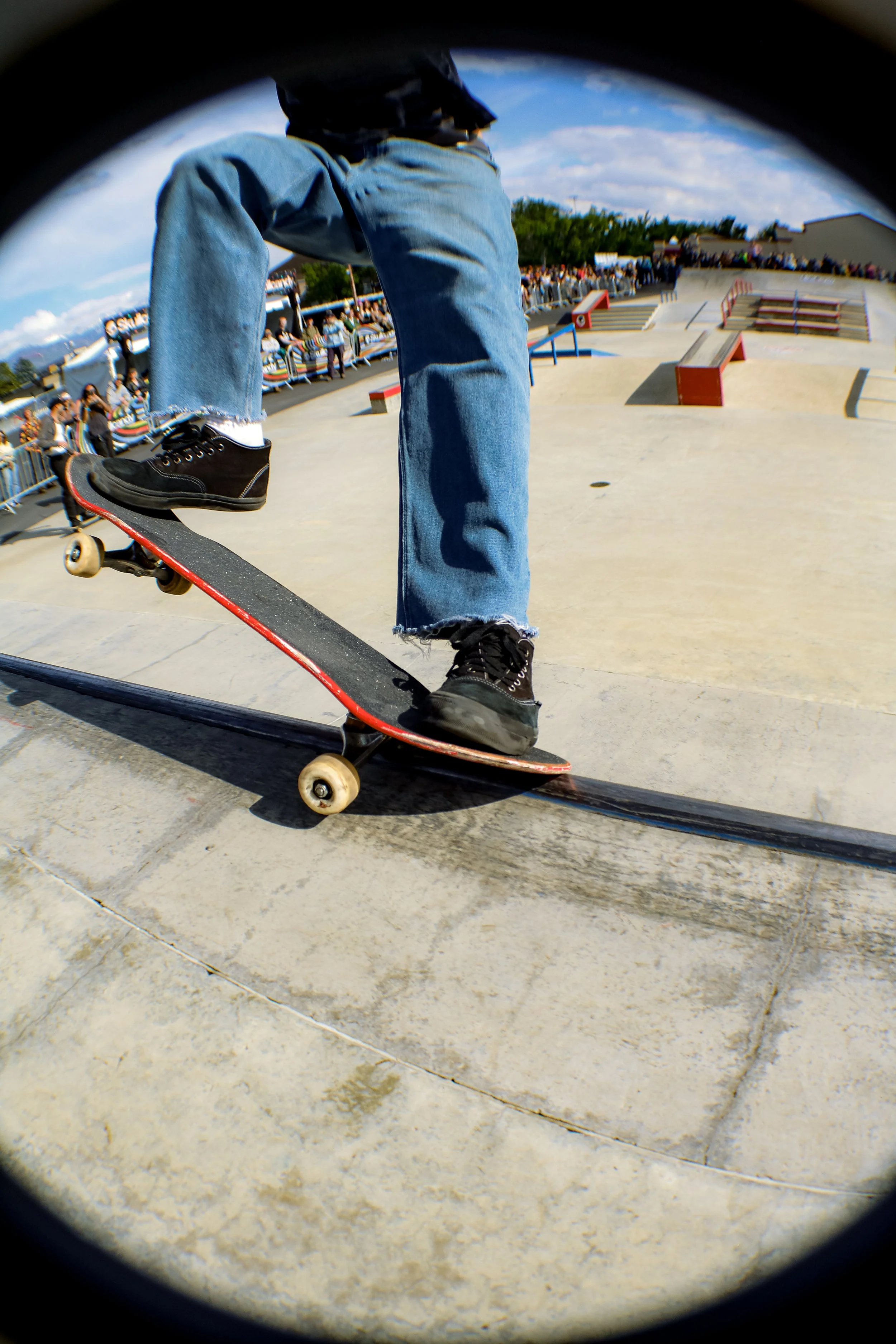 A skateboarder performing a trick at a skatepark during daytime, with spectators and skate ramps in the background.