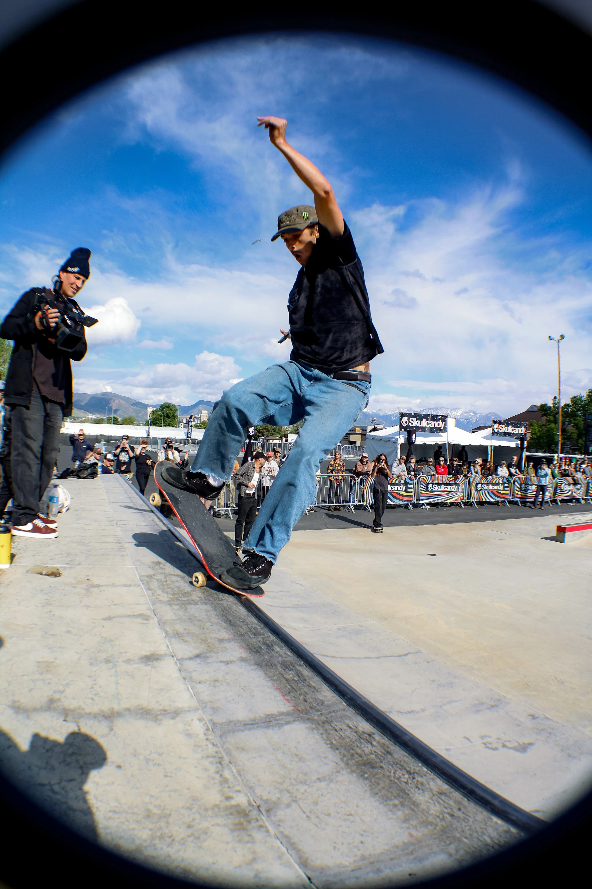 Skateboarder performing a trick at a skatepark with spectators and event tents in the background, under partly cloudy skies.