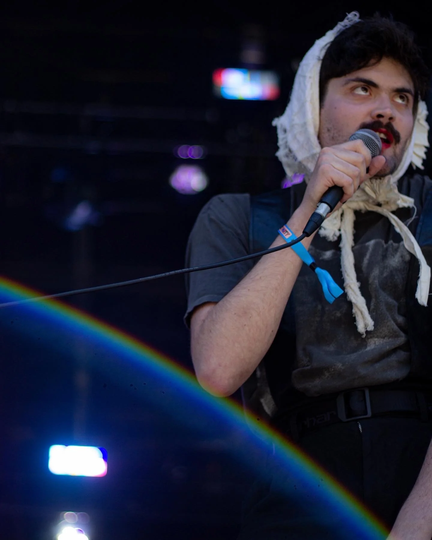 Person holding a microphone, wearing a scarf on their head, on stage with dark background and colorful lighting