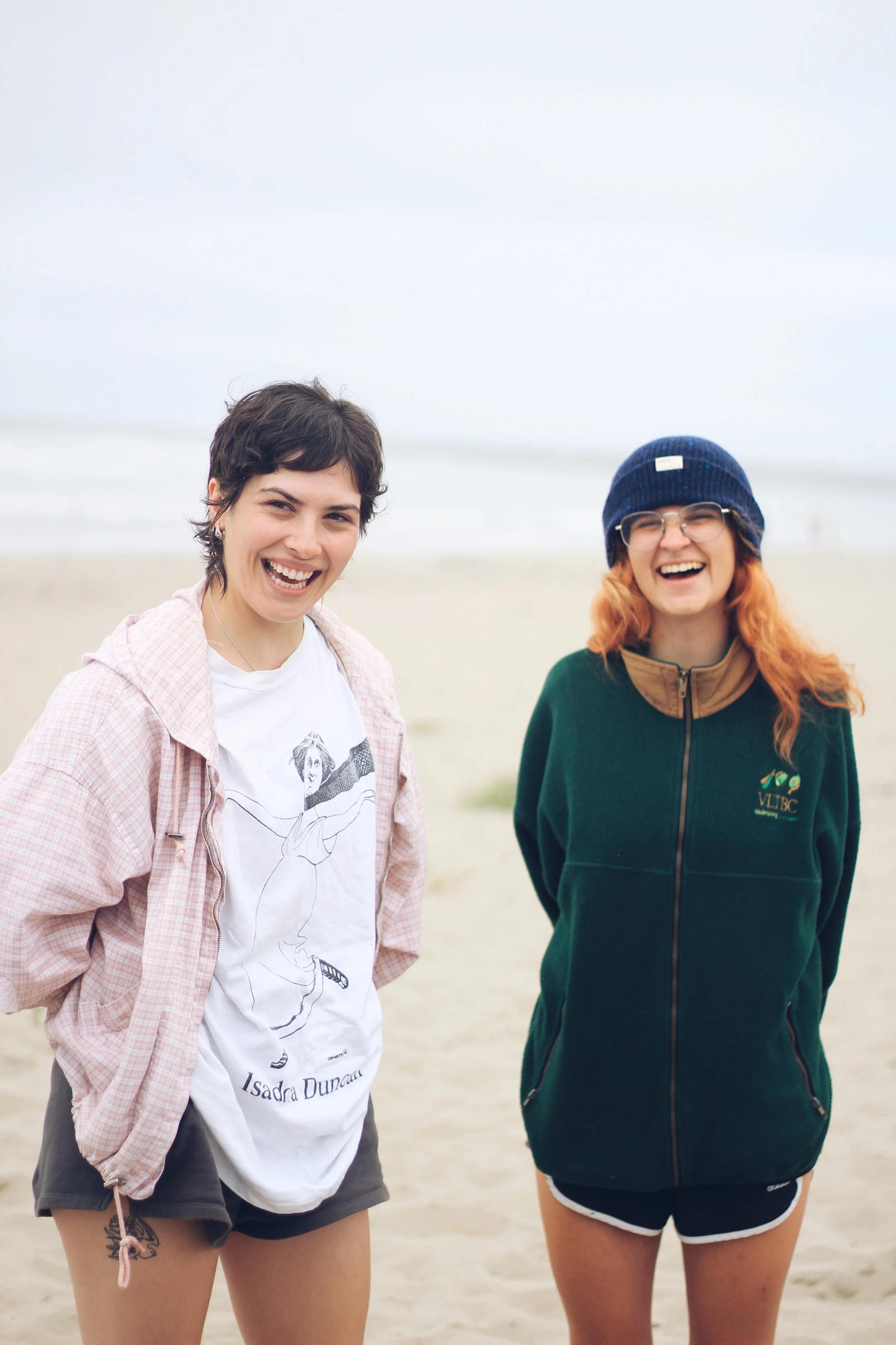 Two young women smiling and standing on a beach with overcast sky in the background.