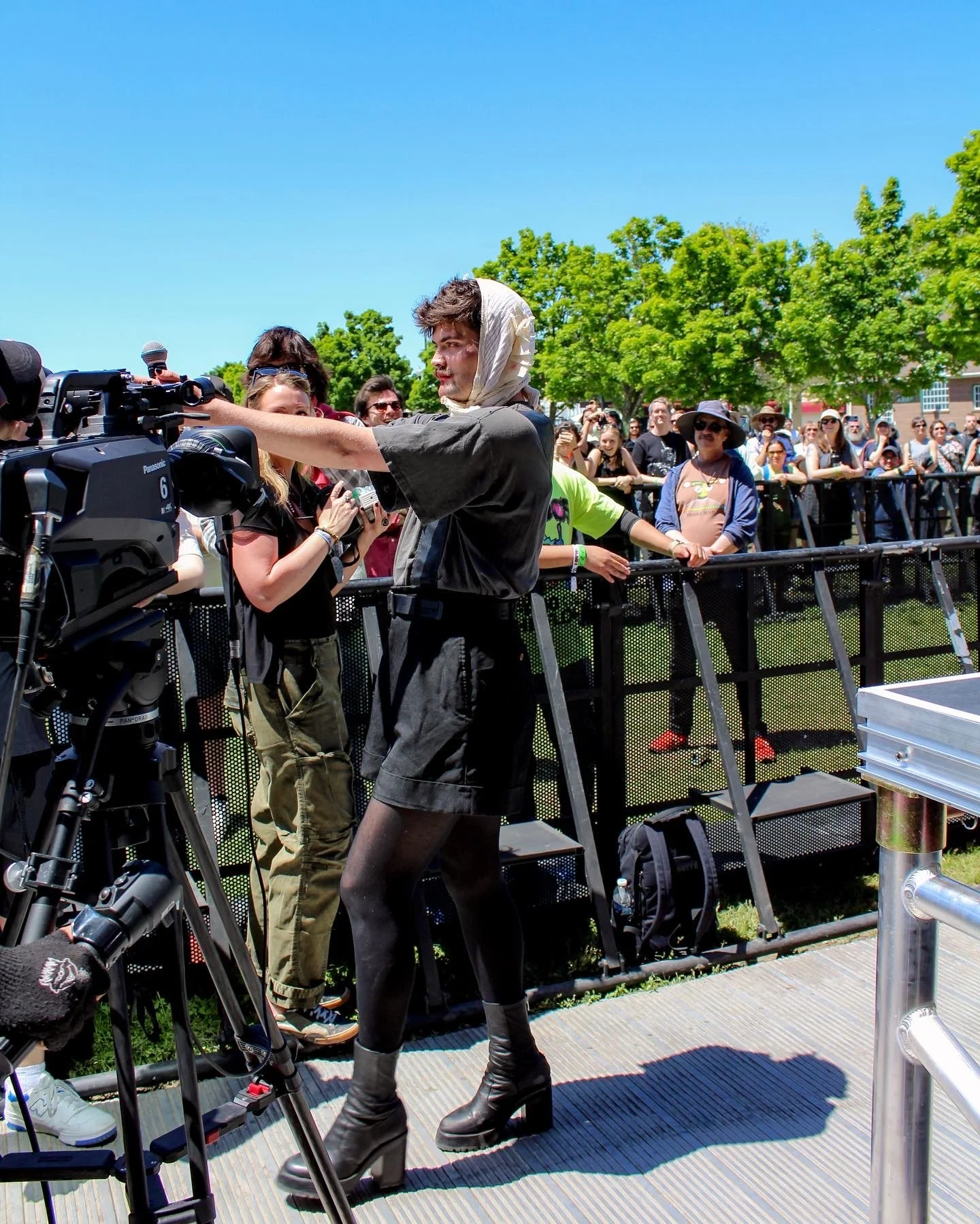 A young woman with a white scarf around her head, wearing black tights, a black shirt, and platform boots, stands in front of a camera at an outdoor event, with a crowd of people behind a barrier on a sunny day with green trees.