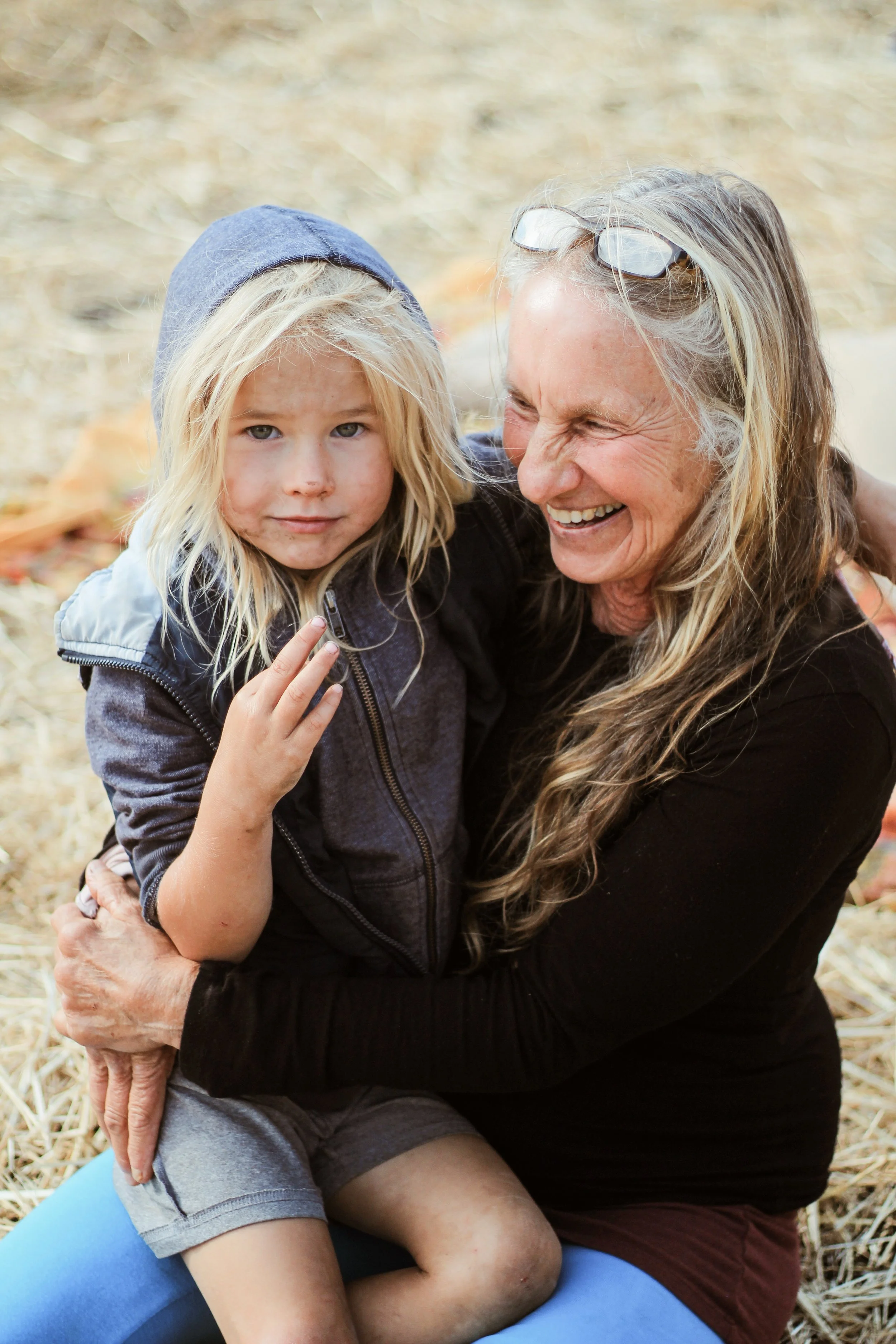 A young girl with long blond hair and a woman with long, grayish hair are sitting outdoors on the ground covered in hay. The girl wears a blue hoodie and gray shorts, and she has dirt on her face. The woman is smiling and holding the girl in an embrace, wearing glasses on her head and a black top. The background is blurred hay or straw.