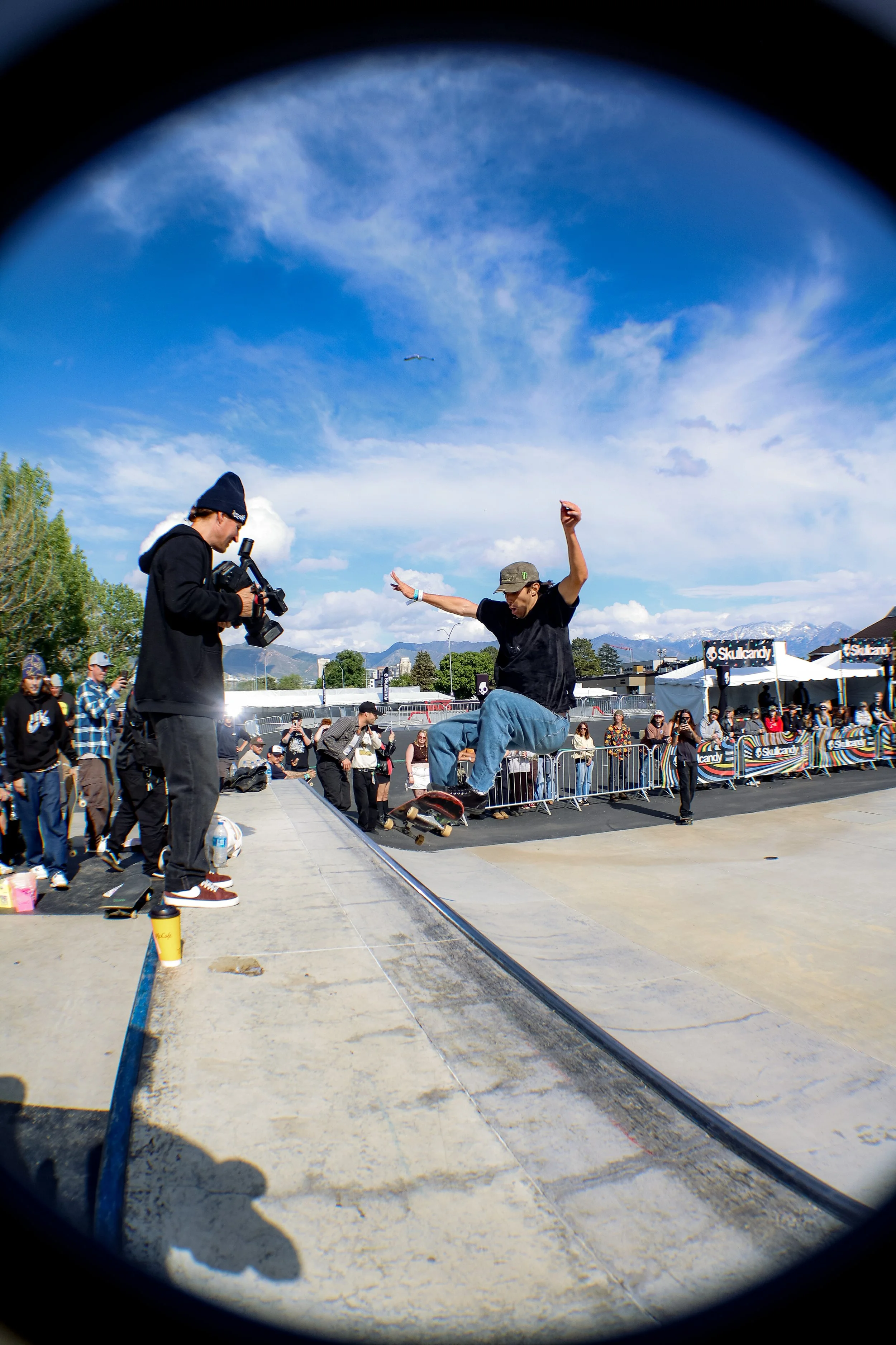 Skateboarder mid-air at an outdoor skate park, with onlookers and a cameraman capturing the moment, under a blue sky with clouds.