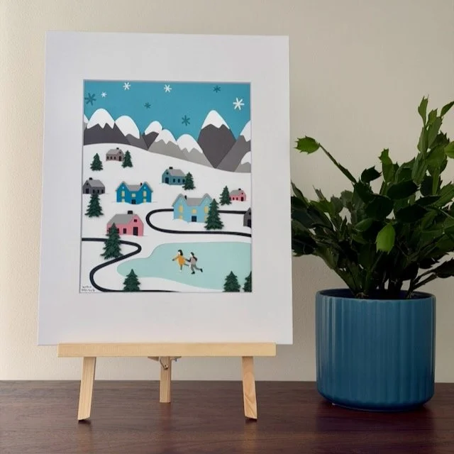 Winter Wonderland Village original paper illustration on a small easel on a wood shelf beside a green plant