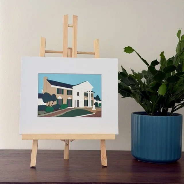 Graceland original paper illustration on a small easel on a black metal shelf beside a green plant
