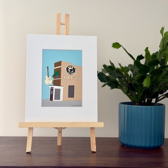 Sun Studio original paper illustration on a small easel on a wood shelf beside a green plant