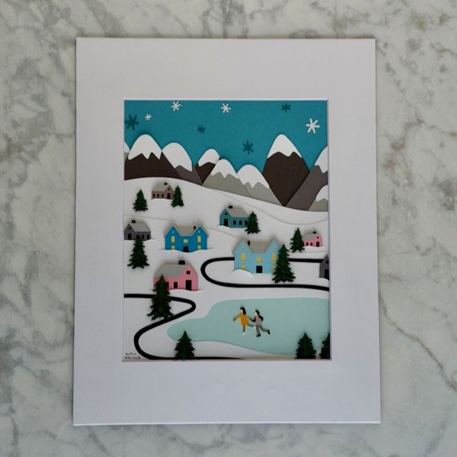 Winter Wonderland Village original layered paper illustration in a white mat, snowy village with evergreen trees and two ice skaters