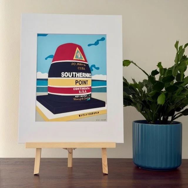 Southernmost Point original paper illustration on a small easel on a wood shelf beside a green plant
