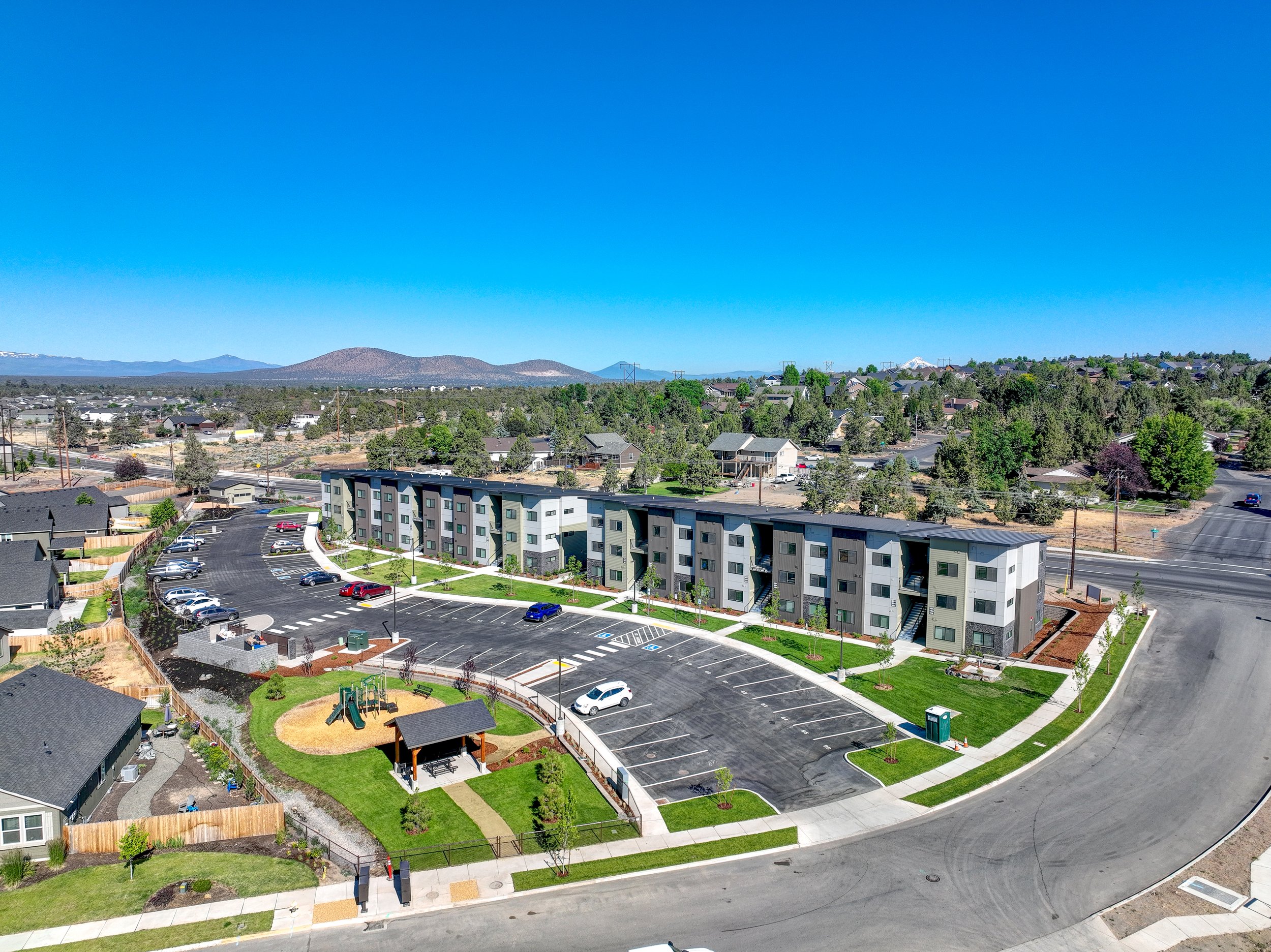 Aerial view of a residential apartment complex with surrounding parking lot, playground, and landscaped area, situated in a suburban neighborhood with mountainous backdrop.