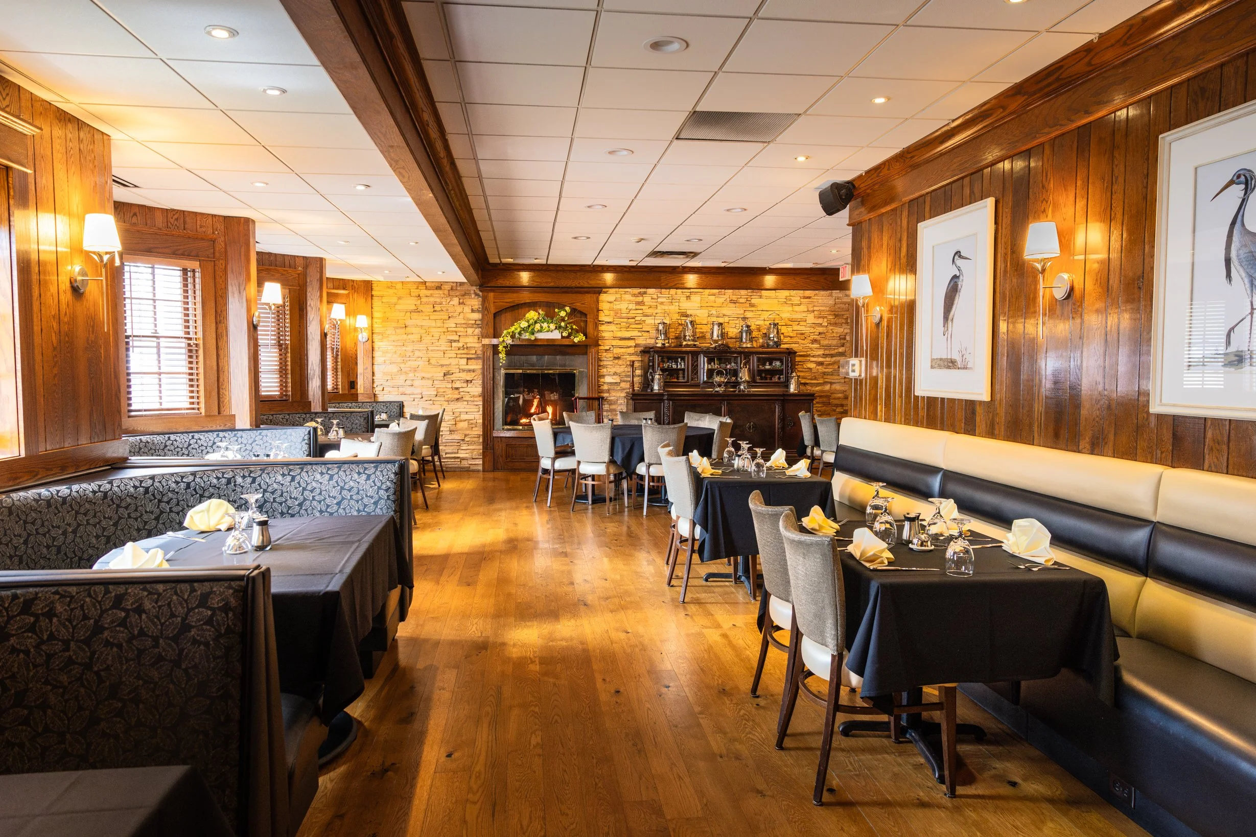 Dining room with wooden walls, hardwood flooring, tables with black tablecloths, and beige chairs. Wall art with bird illustrations, fireplace, and decorative items visible.