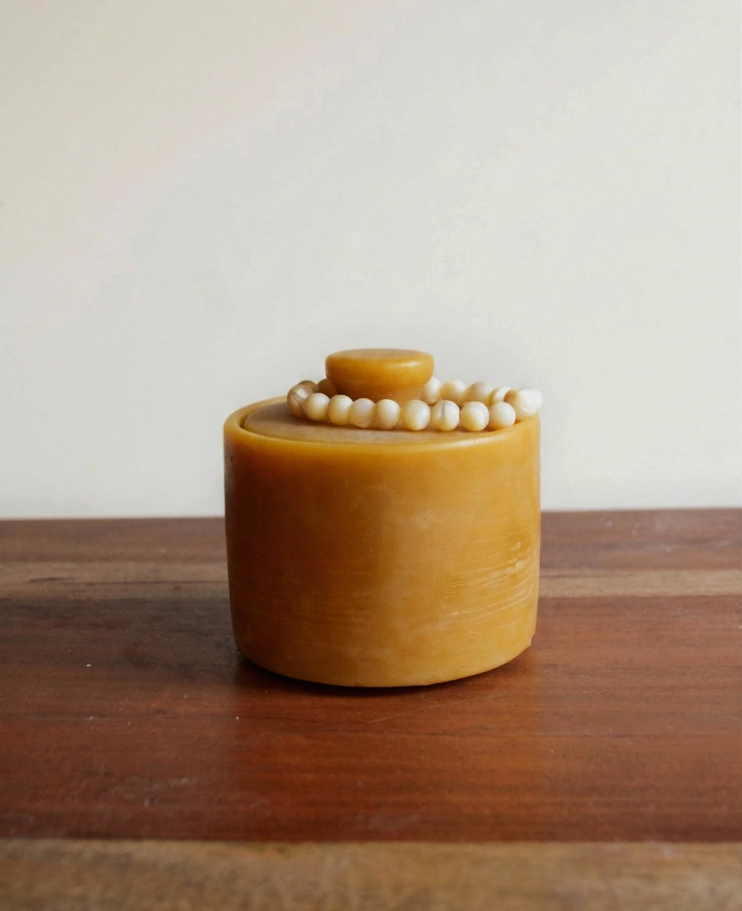 A small, round, orange-brown container with a lid, decorated with a string of white beads, placed on a wooden surface with a plain white background.