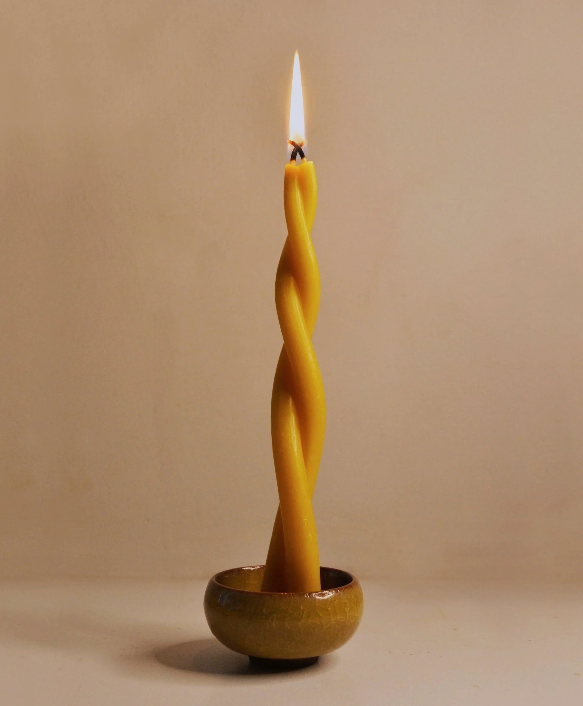 A handmade beeswax twisted tapered candlestick, lit in a small ceramic candleholder. Clean burning unscented natural candlelight.