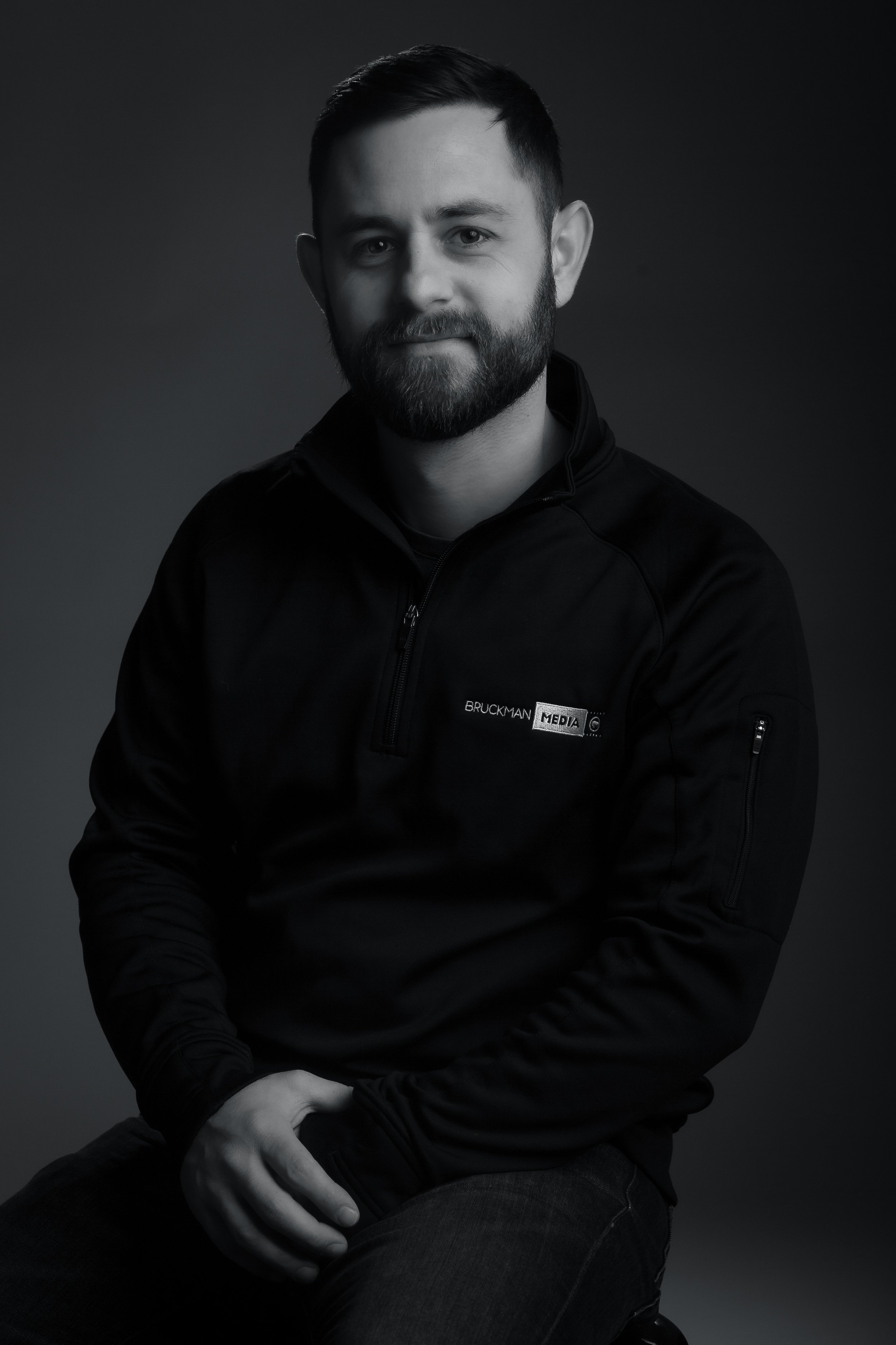 Black and white portrait of a man with a beard wearing a jacket with "Bruckman Media" logo.