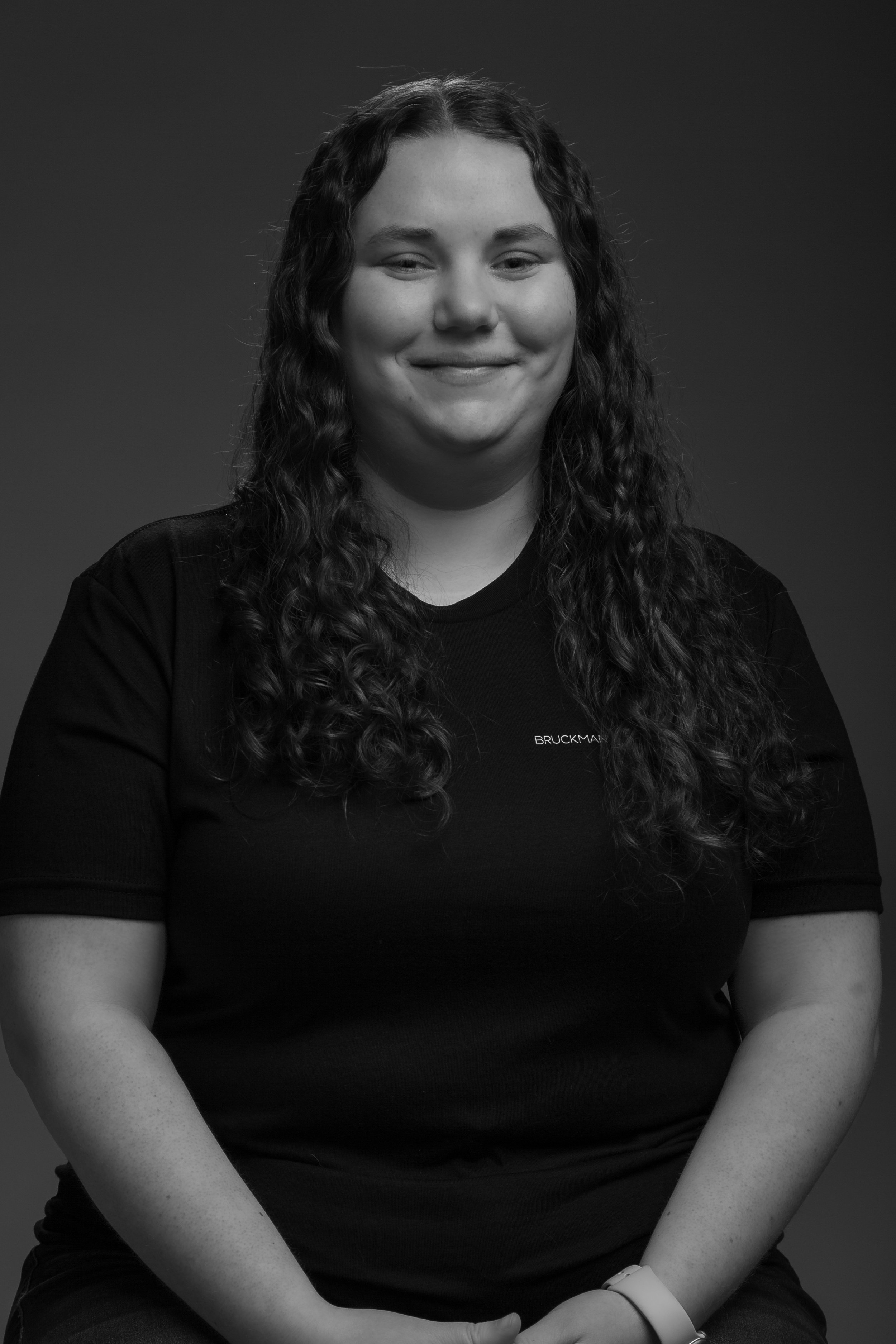 A black and white portrait of a woman, Sam, with long, curly hair, smiling slightly, wearing a dark shirt with a Bruckman Media logo on it, and a watch on her left wrist.