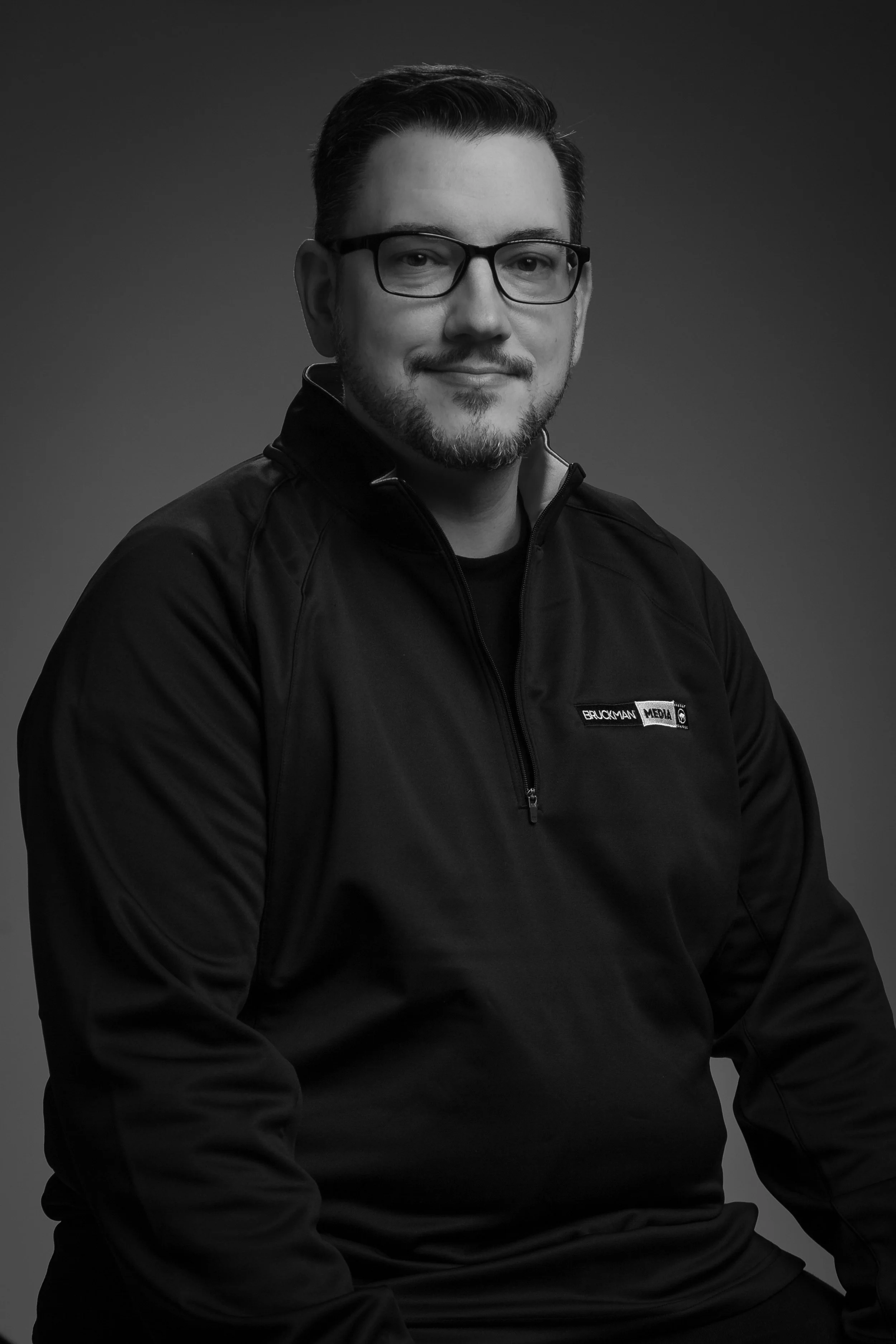 A black and white portrait of a man, Pat, with glasses, beard, and mustache, wearing a jacket with a Bruckman Media logo on the chest, smiling softly.