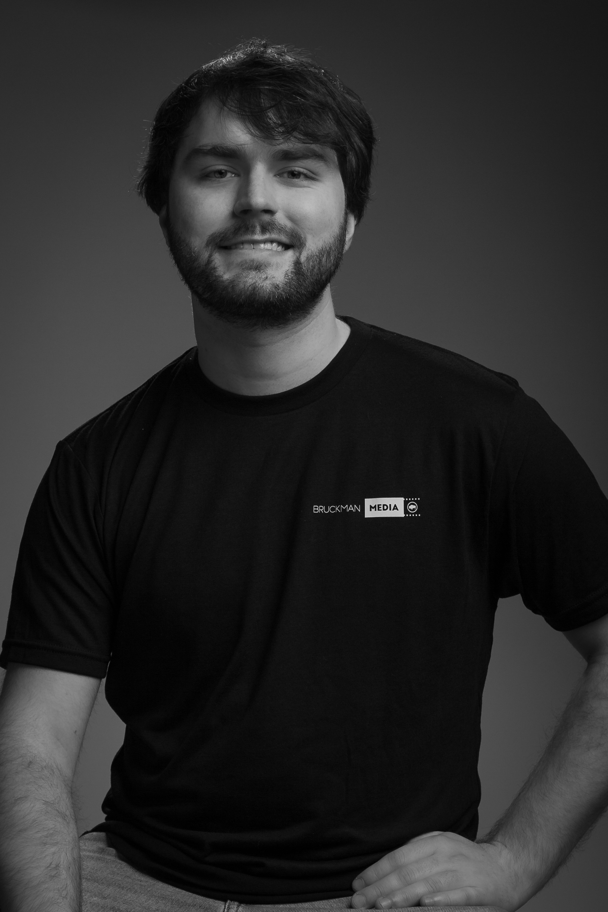 Black and white photo of a young man, Ted, with a beard, wearing a black T-shirt with the words 'BRUCKMAN MEDIA' on it, smiling slightly, with his right hand resting on his lap, against a dark gradient background.