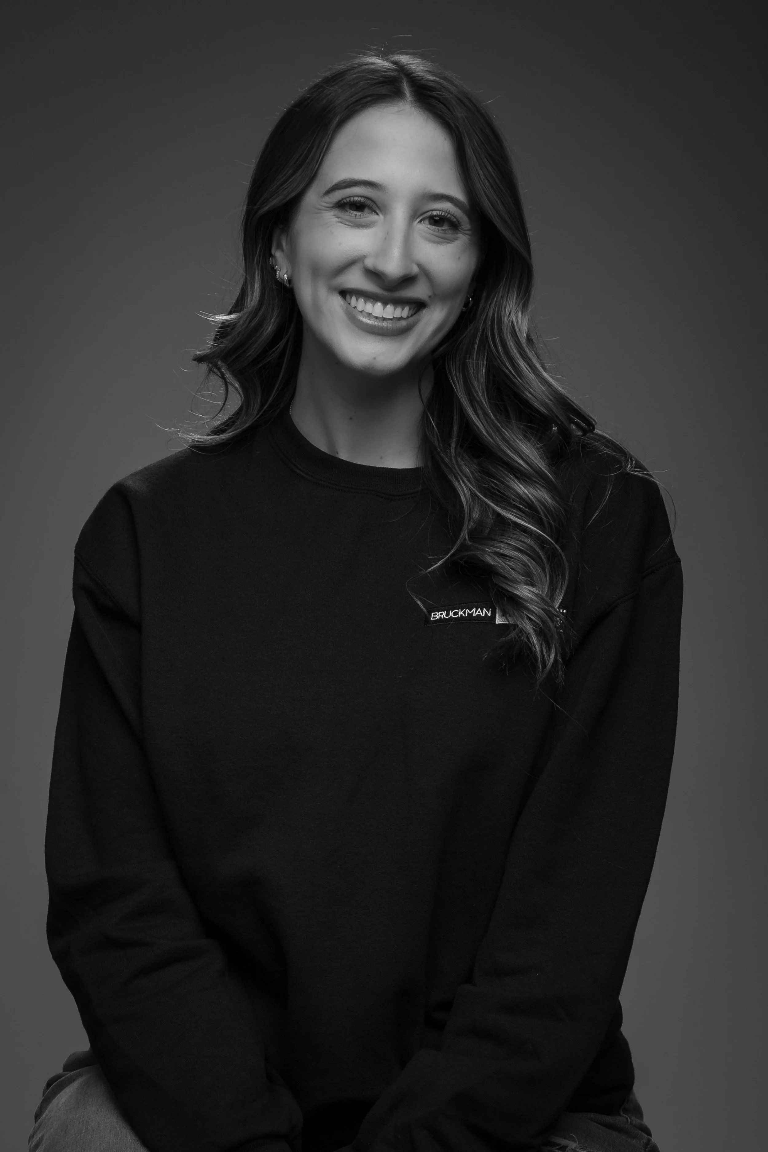 Black and white photo of a smiling woman, Liz, with long wavy hair and earrings, wearing a dark sweatshirt with a small Bruckman Media logo on the chest, against a plain background.