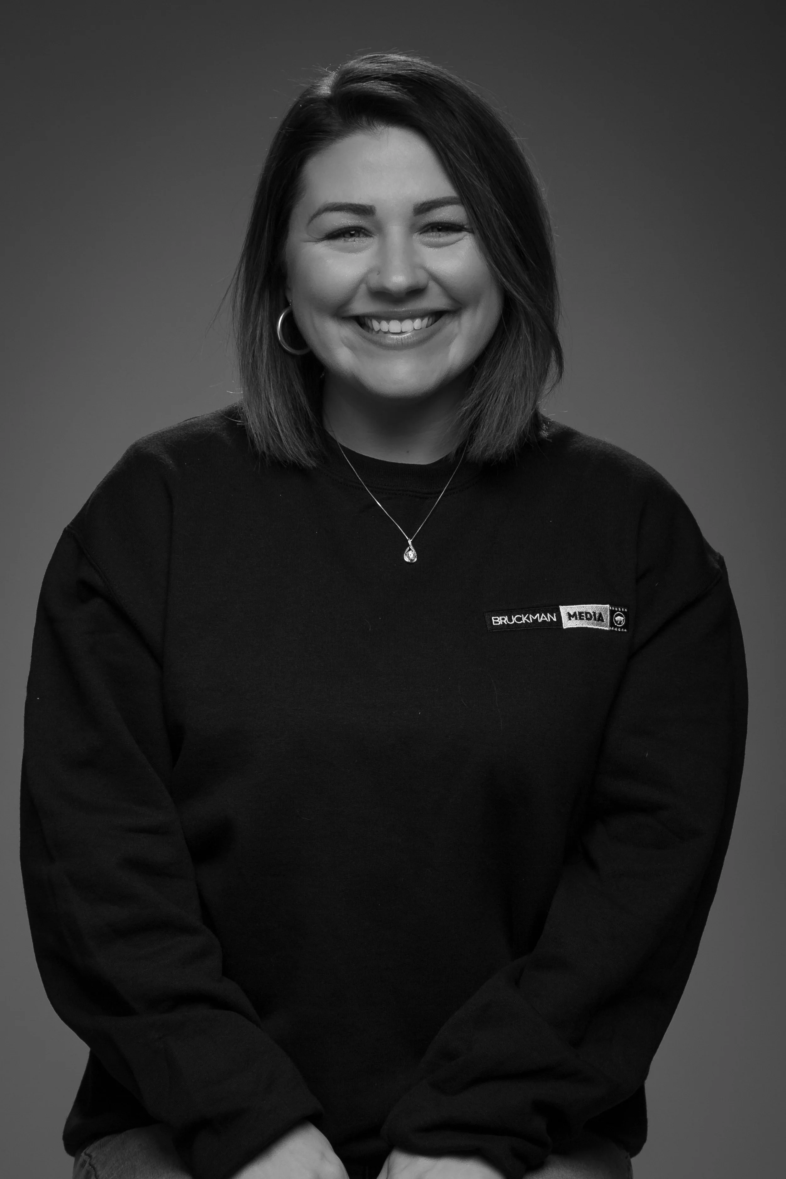 A young woman, Jess, with shoulder-length hair smiling, wearing hoop earrings, a necklace, and a black sweatshirt with Bruckman Media logo, posed against a grey background.