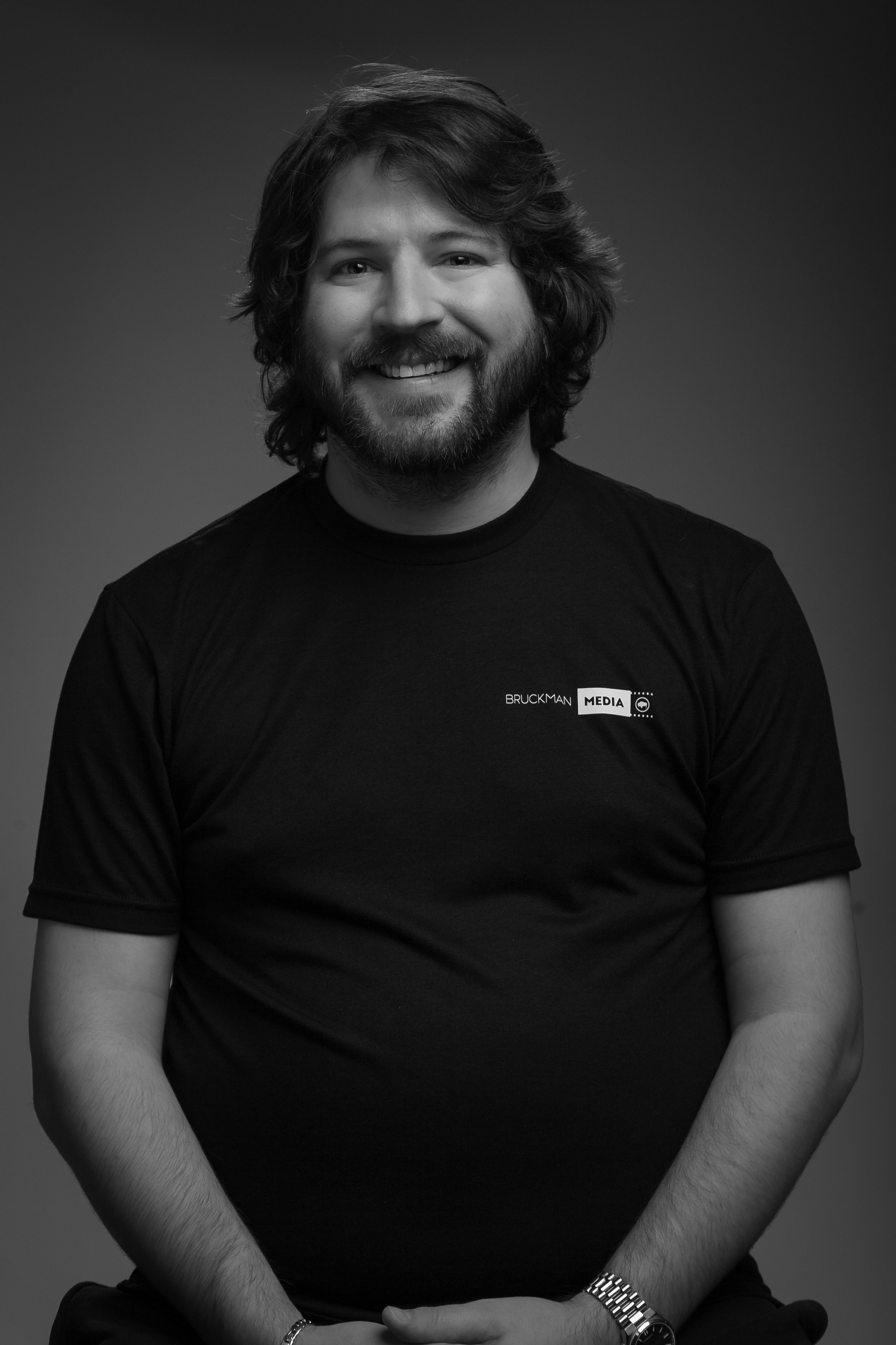 Black and white portrait of a man, Donny, with long hair and a beard, smiling, wearing a black T-shirt with a Bruckman Media logo, sitting against a plain background.