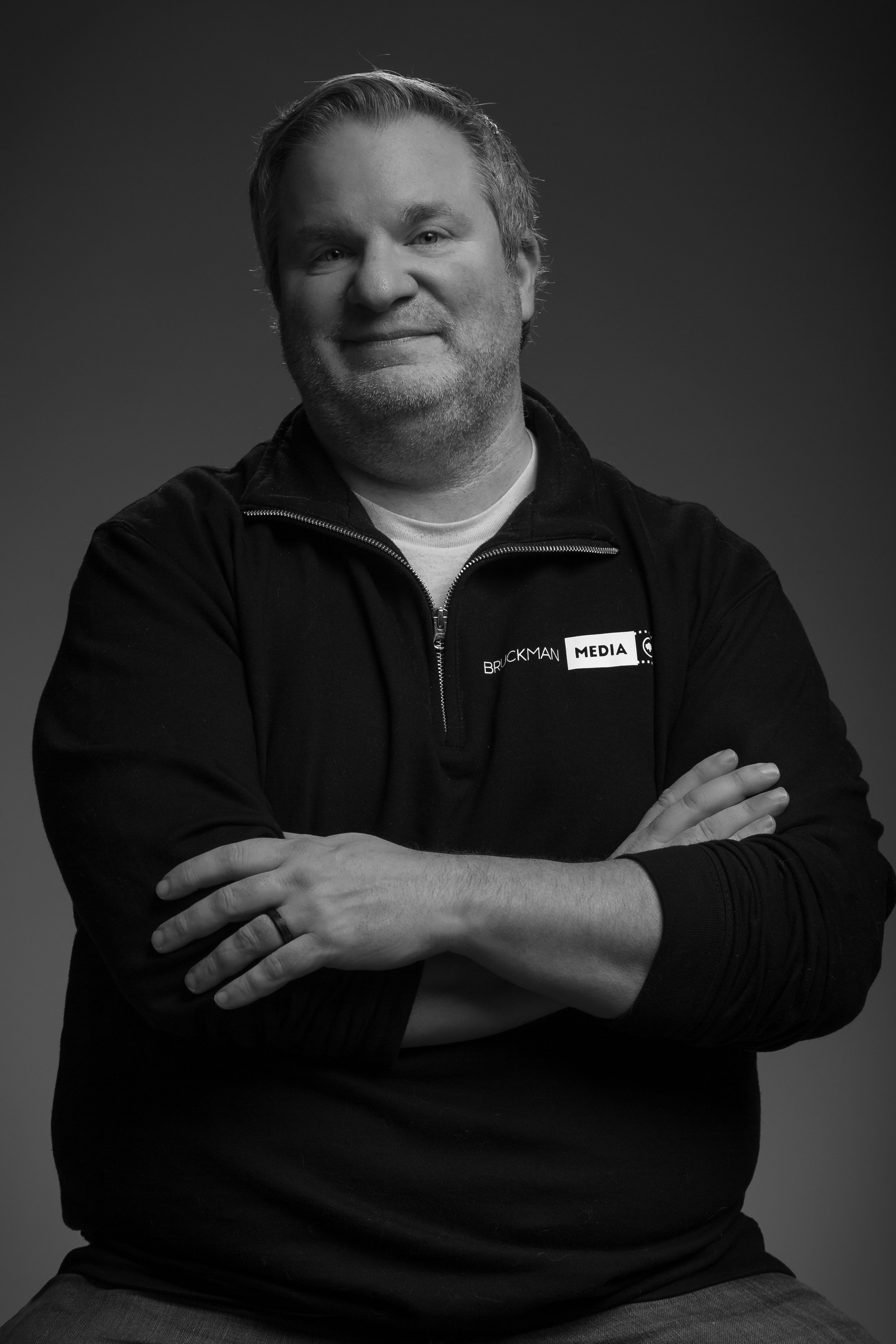 A black and white portrait of Bill, a man with short hair and a beard, crossing his arms in front of him, wearing a zip-up sweatshirt with a logo that says "BRUCKMAN MEDIA".