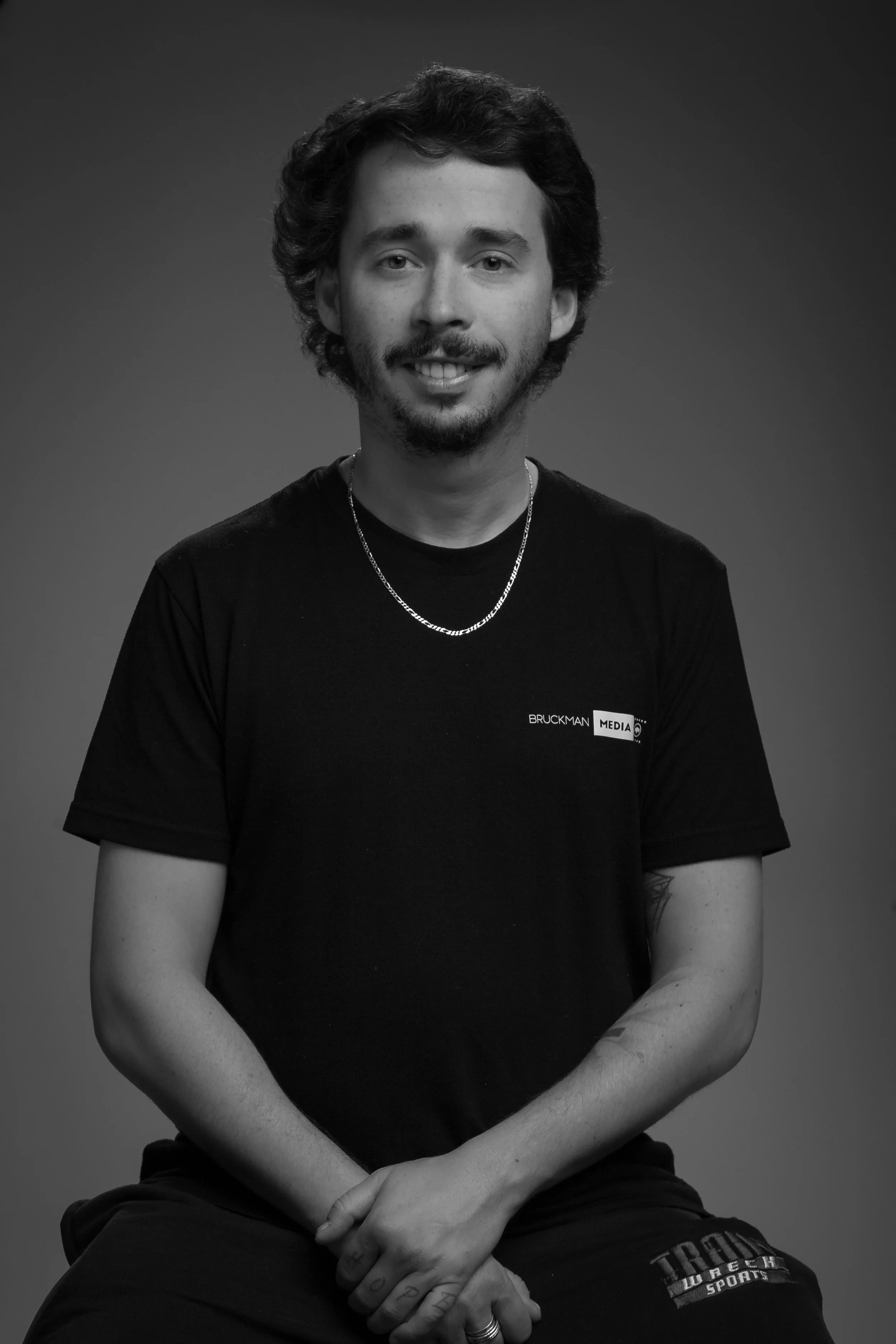 Black and white portrait of a young man, Elliot, with curly hair, a light beard, wearing a T-shirt with a Bruckman Media logo, a chain necklace, and a ring on his finger, sitting against a plain background.