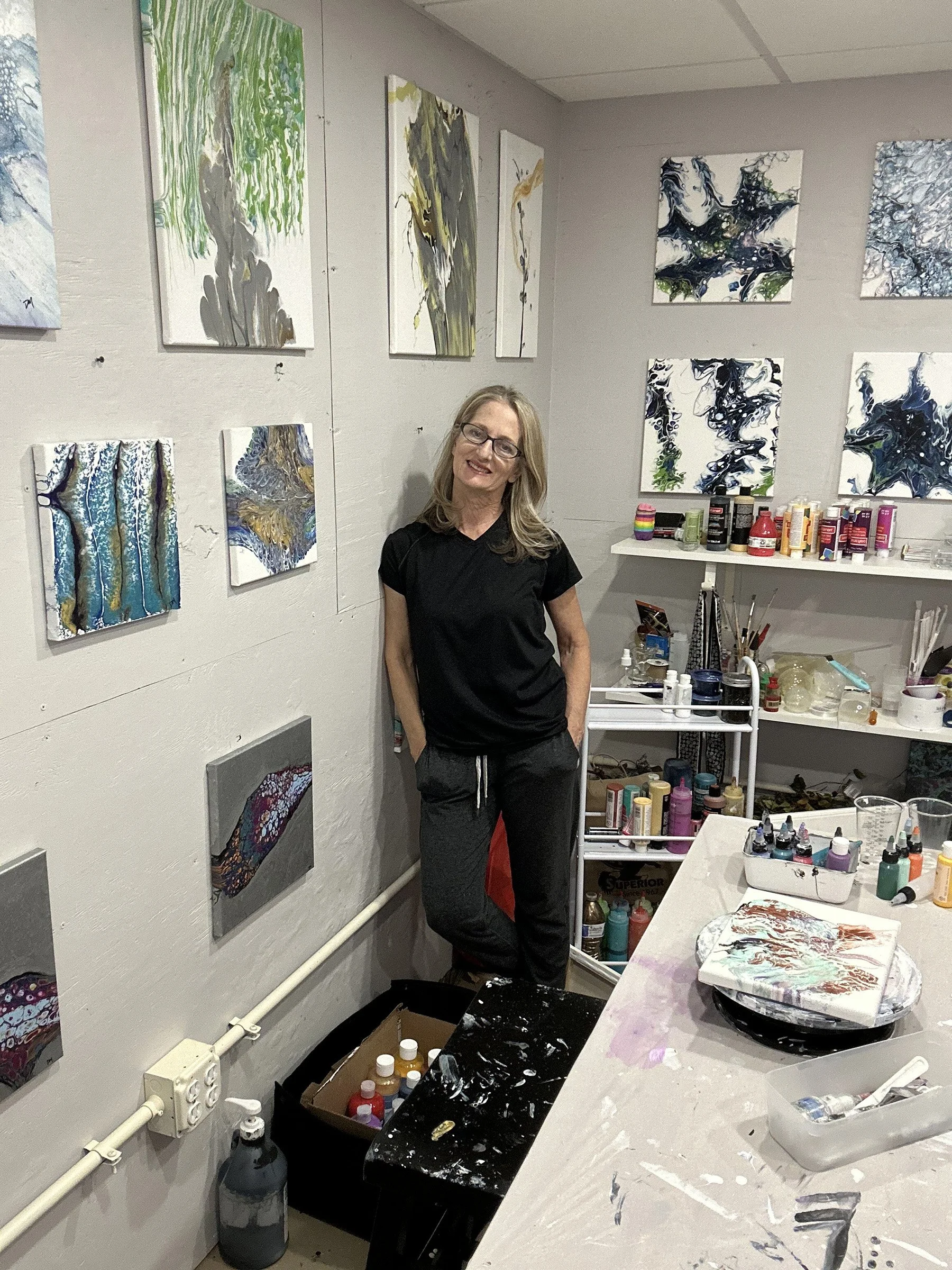 An art studio with abstract paintings on the wall, a woman wearing a black shirt and glasses standing next to the artwork, and a table with painting supplies and bottles of paint.
