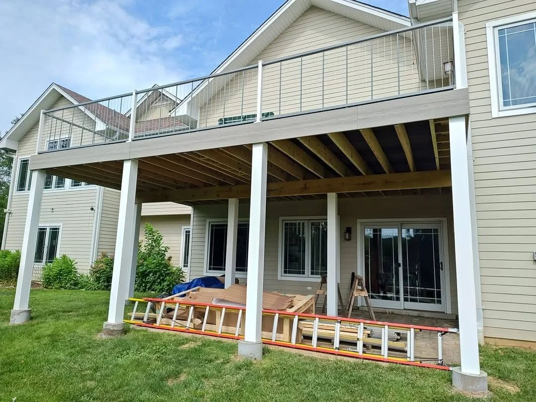 Back of a house with ongoing deck construction, showing the under-assembly of a new upper deck with white railing and support beams, construction materials and tools on the ground.