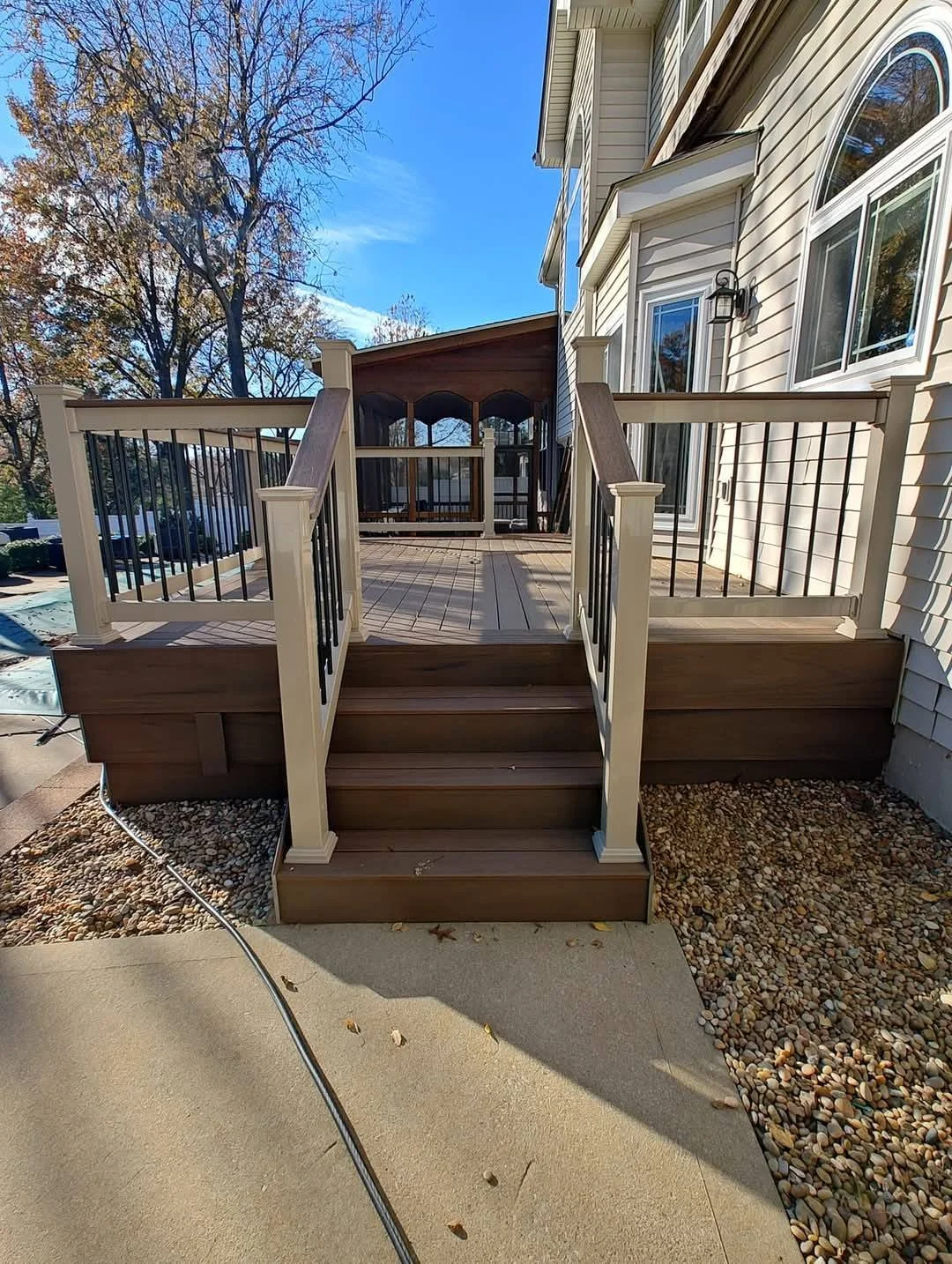 Wooden deck with stairs, beige and black railings, attached to the white house, with trees in the background and a blue sky.