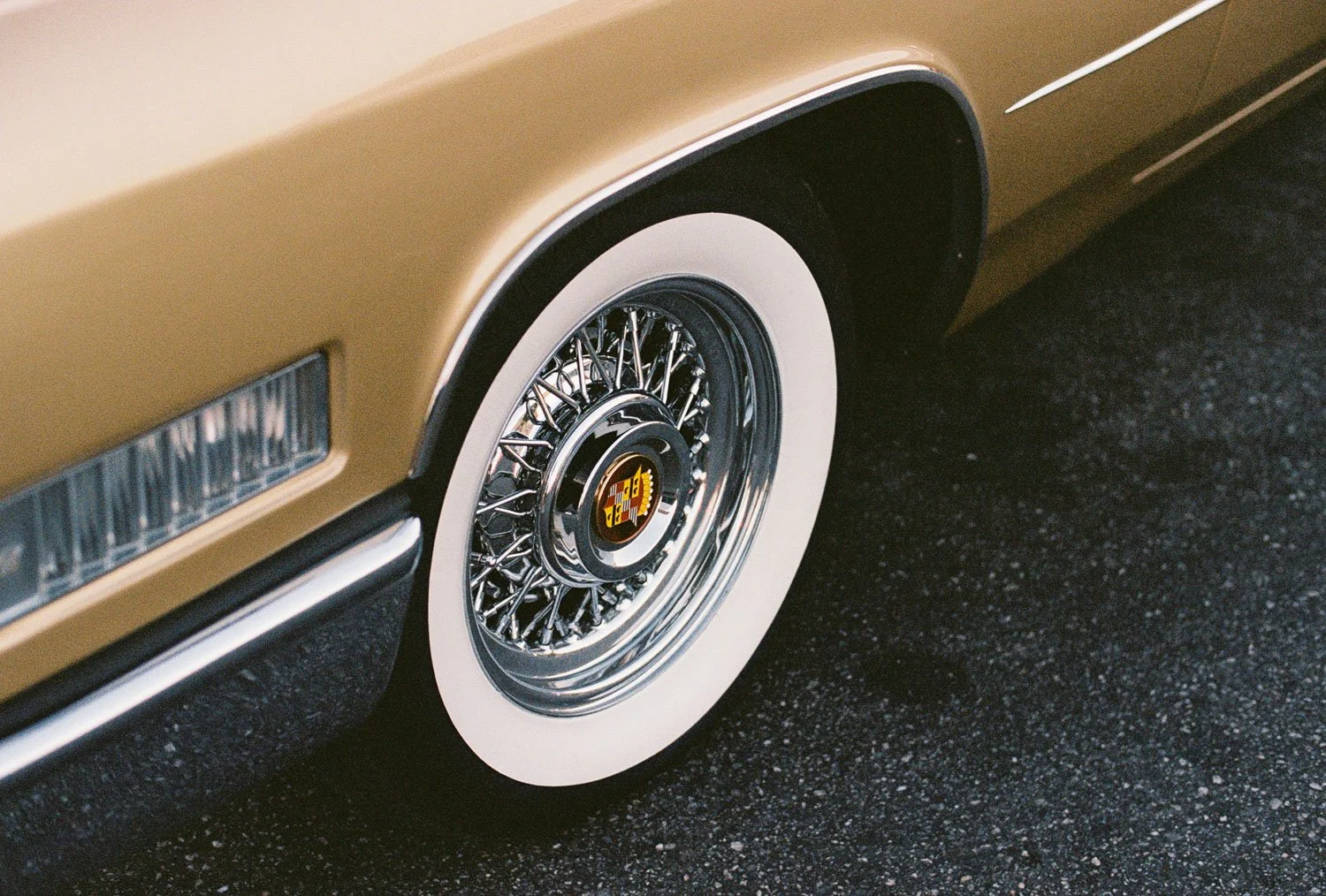Close-up of a vintage gold-colored car wheel with whitewall tire and wire spokes, featuring a hubcap with a colorful logo