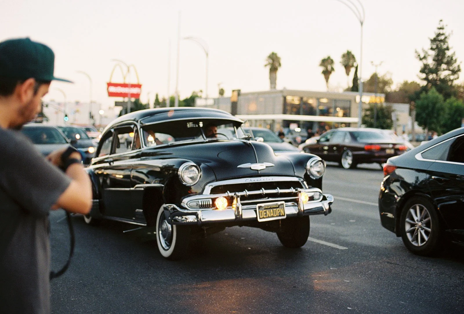 A vintage black Chevrolet car with a customized license plate reading 'DENADPN' is parked on a busy city street during the evening, alongside modern vehicles, with palm trees and a McDonald's sign in the background.