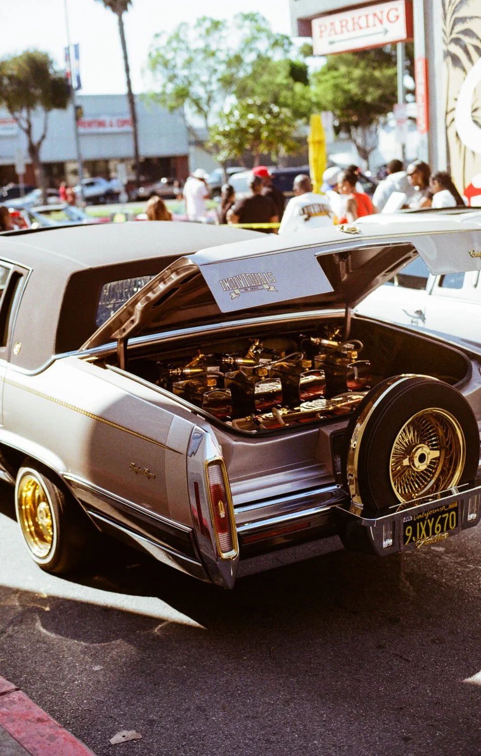 Rear view of a classic car with its trunk open, showing a collection of gold-colored rims and polished tools, parked at an outdoor event with people and trees in the background.